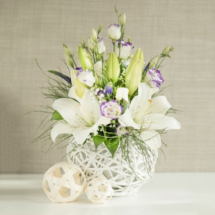 A tranquil floral centrepiece in shades of white, cream, and lilac is shown resting on a pure white surface, the kind you might see decorating a wedding table in a Mill Hill marquee. The arrangement is set in a round white vase with a striking open-weave, basket-like texture, its airy lattice catching the light and lending a stylish yet natural character. From this base, several white lilies extend upward, some blooms fully open with elegant, flared petals and clearly visible stamens, while others remain closed as elongated, pastel green buds, adding height and freshness.
Interwoven among the lilies are delicate lisianthus flowers, their soft white petals edged or kissed with subtle lavender-purple tones. A mix of buds and blossoms provides a pleasing rhythm, with tightly curled buds giving way to layered, rose-like blooms. Tiny white filler flowers, reminiscent of baby's breath, are dusted throughout the display, and a few deeper violet accents hide between the stems, giving a touch of richness to the light colour palette. The arrangement is supported by generous greenery: feathery fronds, perhaps similar to asparagus fern or soft ruscus, mingle with broader leaves to provide both texture and volume. Several stems of foliage drape slightly over the rim of the vase, softening its silhouette and lending a natural, informal grace.
In front of the vase sit two decorative spheres, crafted from a pale woven material that echoes the vase's open lattice. The background shows a light beige wall with subtle horizontal lines, as if covered in fine linen, gently illuminated so the whites glow warmly. The result is a peaceful and sophisticated display, fitting for weddings, moments of remembrance, or refined home décor across Mill Hill and neighbouring Green Belt streets.