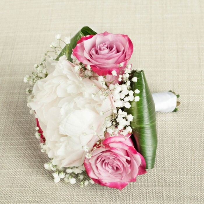 Gently placed on a fine, textured fabric reminiscent of Mill Hill's elegant interiors, this sophisticated bouquet captures the artistry and thoughtful detail that define Flowers Mill Hill. At its heart, two luminous pink roses-petals deepening from rich fuchsia at the edges to soft, inviting hues of blush and lilac in their swirling centers-anchor the design at opposing ends, guiding the gaze toward a voluminous white peony in full bloom. The peony's dense ruffles of creamy petals create an inviting, cloud-like effect, drawing in the gentle light and offering an impression of pure abundance. An airy scattering of white baby's breath is woven throughout, elevating the bouquet's softness and filling every small gap with ethereal, starry blossoms. The lower portion is elegantly collared by broad, deep green foliage, likely ti or aspidistra leaves, folded and structured to provide a modern yet natural finish. The stems are tightly gathered, wrapped in a pristine white satin ribbon that forms a classic, hand-held bouquet-ready for a Mill Hill bride to carry down the aisle or a loved one to receive on a momentous day. The neutral, woven background ensures the vivid pinks, soft whites, and glossy greens stand out, while the composition's balanced lighting brings every detail to life. This arrangement would lend a romantic flourish to events at Mill Hill Synagogue, engagement parties along Flower Lane, or any elegant occasion across NW7.