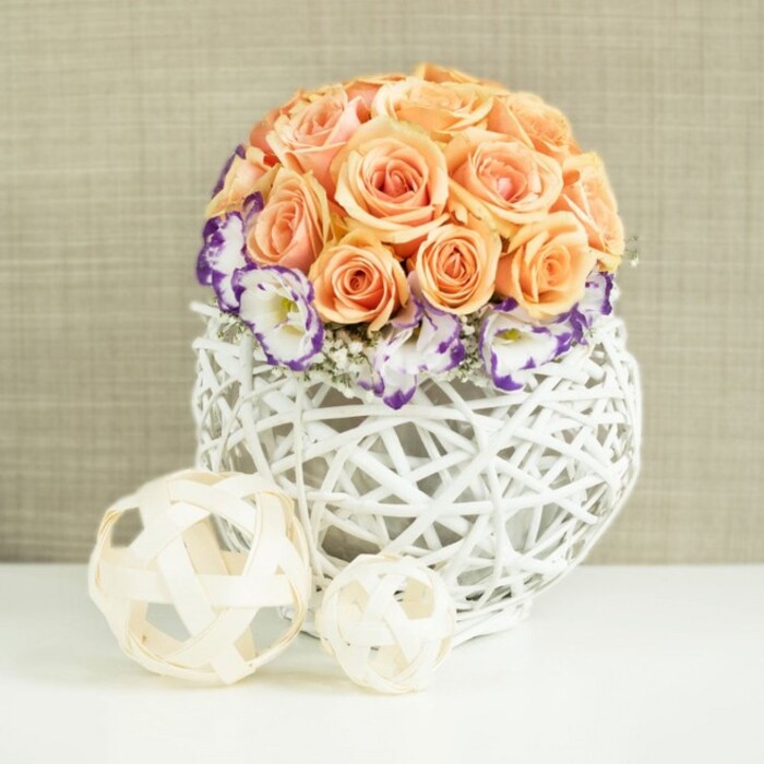 Set against a softly textured beige backdrop, a romantic floral centerpiece takes pride of place on a pristine white surface, the sort of arrangement you might see in a beautifully dressed Mill Hill dining room on a special evening. At its core sits a perfectly domed cluster of roses in mellow peach and apricot shades, with hints of soft salmon and blush. The roses are densely packed, their velvety petals layered in tight spirals that curl outward, so the dome appears full, luxurious, and exceptionally neat. Encircling the base of this rose canopy is a band of lisianthus, their creamy white petals ruffled and finely edged with a deep violet or purple outline, creating a sophisticated contrast that frames the warmer tones above. Among these, small sprays of white baby's breath weave in and out, their tiny star-like flowers adding a delicate, cloudlike texture that lightens the whole composition. The arrangement is nestled into a distinctive white woven container, with a broad, bulbous shape reminiscent of a modern basket. Its interlaced wicker strands cross and twist to form an airy lattice, allowing glimpses of shadow and the stems within, providing a blend of rustic craft and contemporary style that would feel at home in a Mill Hill flat or a venue just off Lawrence Street. In front of the vase, two decorative spheres rest on the white surface, one larger than the other, created from thin, pale wooden strips woven into open, spherical forms that subtly echo the container's structure. The neutral fabric background, with its gentle, linen-like texture, adds warmth and depth without drawing attention from the flowers. Together, the elements create an atmosphere of quiet celebration and thoughtful design, ideal for weddings, anniversaries, or refined table décor in Mill Hill.