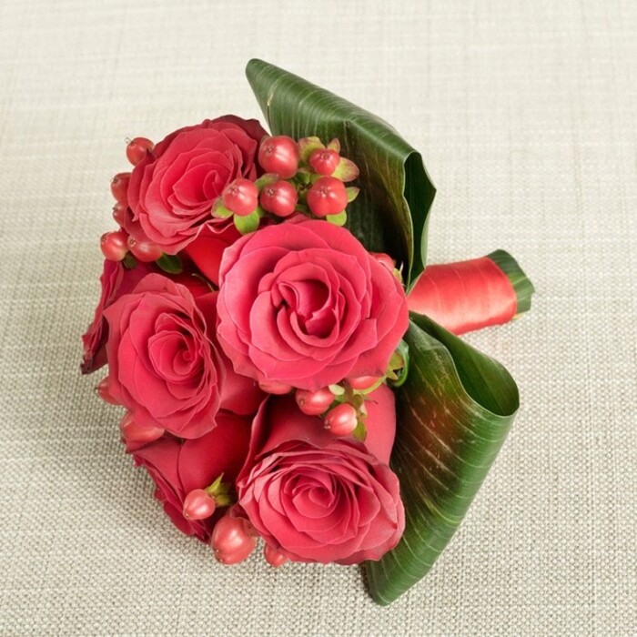 A richly romantic bouquet of deep red roses and berries is displayed on a light beige, finely textured fabric, the neutral background gently echoing the calm interiors of homes around Mill Hill while allowing the vivid blooms to command attention. The arrangement is tightly composed, with around six fully opened roses gathered into a low, rounded cluster, their velvety petals layered densely in spirals of crimson and scarlet. Some petals curl inwards, almost black-red at their depths, while others open outward in wider arcs, giving a sense of fullness and life, as though the roses have just reached their perfect moment of bloom. Nestled between these roses are dainty clusters of Hypericum berries in rich shades of dark red and rosy pink, each small, bead-like fruit smooth and slightly glossy, adding a playful, almost jewel-like sparkle and gentle texture contrast to the softness of the petals. Embracing the bouquet, broad, deep green tropical leaves are skillfully curved and wrapped around one side, their pronounced veins and polished surface creating a strong, sculptural frame. Around the stems, mostly tucked beneath the flowers but still noticeable, a luxurious red satin ribbon is tied snugly, its sheen catching the light in a way that matches the opulence of the roses. The fabric beneath is a soft cream-beige, with a subtle weave that reads as sophisticated yet understated, helping the rich reds and greens to pop without distraction. The overhead, slightly angled viewpoint and gentle, even lighting evoke a quiet, intimate atmosphere, suggesting a hand-tied gift prepared for a Mill Hill anniversary, a Valentine's surprise delivered near Mill Hill Park, or a refined wedding accent created with expert floristry skills and a deep understanding of romantic design.