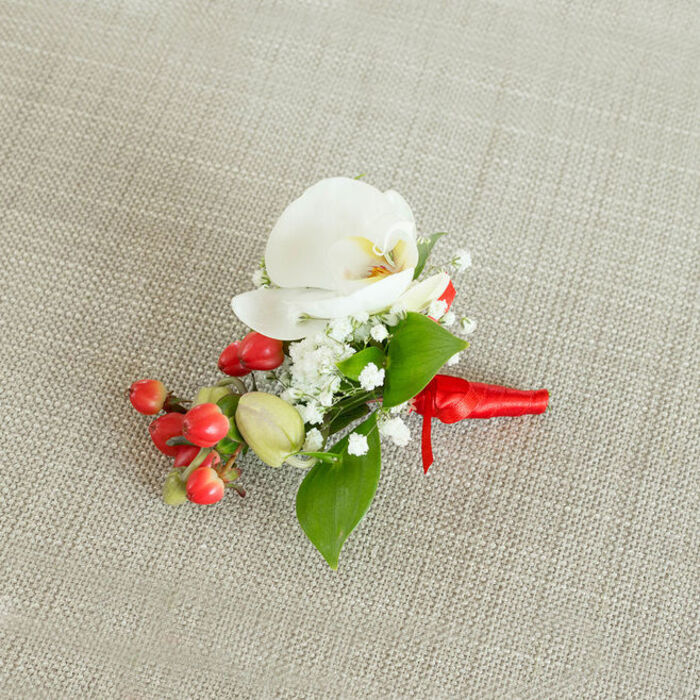 This image presents a refined floral boutonniere lying on a softly textured, light beige fabric that resembles natural linen, the kind often used in intimate Mill Hill wedding receptions. Photographed from a slightly raised overhead angle, the composition allows every detail of the piece to be clearly seen. At the centre is a striking white Phalaenopsis orchid, its wide, gently curved petals opening in a graceful arc. The orchid's throat carries a subtle mix of creamy yellow and soft orange, adding warmth and definition to the otherwise pure white bloom and drawing the eye into its heart. Surrounding the orchid are sprays of dainty baby's breath, each stem topped with clusters of tiny white florets that create a gauzy, cloud-like effect. These delicate blossoms soften the strong lines of the orchid and add a romantic haze of texture. Alongside them sits a rounded cluster of bright, cherry-red berries, smooth and glossy, that provide a vivid colour contrast suggestive of winter celebrations and festive gatherings across Mill Hill and Totteridge. A few pale green, oval buds are interspersed among the red berries, hinting at freshness and growth. Frames of polished green leaves, both broader pointed shapes and slimmer elongated ones, weave around the flowers and berries to anchor the design with lush, natural colour. The stems are carefully wrapped in a rich red satin ribbon, its surface catching a soft sheen as it winds tightly and finishes in a neat, understated knot with short tails. The neutral, woven backdrop highlights the interplay of white petals, bright berries, and verdant foliage, giving the arrangement a poised yet inviting character. This boutonniere feels ideal for a groom, groomsman, or prom guest in Mill Hill, adding a refined accent of seasonal charm, elegance, and quiet celebration to a dark suit or dinner jacket.