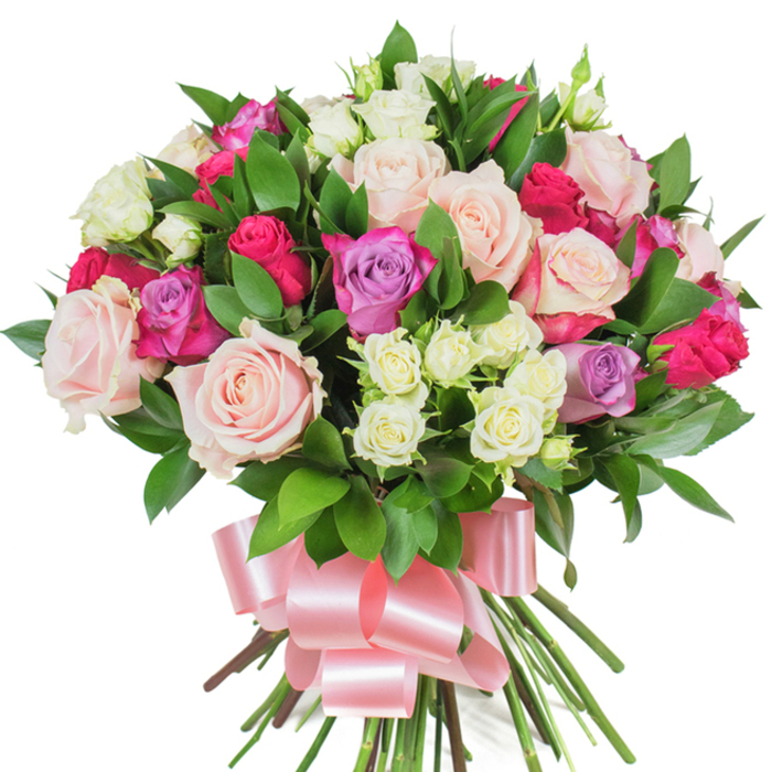 A richly textured bouquet of mixed roses presented against a pristine white background, its dense, rounded form speaking to careful, experienced hand-tying. Prominent are numerous large roses in soft blush pink and creamy white, their layered petals showing delicate veining and a satiny surface that suggests a faint, classic rose perfume when brought close. Among these gentler tones sit striking magenta or deep fuchsia roses that function as joyful colour anchors, while a scattering of lavender roses introduces a pale purple note that softens the arrangement's warmth. Tiny spray roses, mostly pristine white with just a whisper of pale yellow at their centers, nestle into pockets between the larger blooms, offering texture variation and the feel of a bouquet built to be admired up close. Lush, dark green foliage-likely ruscus or salal-frames the blossoms with glossy, elongated leaves that lend both movement and contrast, cradling the florals and making the colours sing. The stems are gathered tightly and finished with a broad, silky baby-pink satin ribbon, tied into an elegant bow whose smooth sheen catches light and echoes the bouquet's romantic mood. Imagined under the soft daylight that filters through a Mill Hill East window or waiting to be placed on a mantel near Bittacy Hill, this arrangement conveys heartfelt sentiment-perfect for anniversaries, Mother's Day, birthdays, or a gracious celebration-delivering a sense of timeless beauty, tactile softness, and considered craftsmanship.