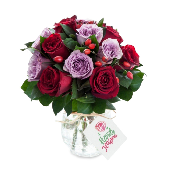 A richly coloured, expertly arranged bouquet showing a balanced duet of deep red and soft lavender roses, crafted with the kind of attention to detail you'd expect from a Mill Hill flower studio. The rounded, dense arrangement alternates velvety crimson and burgundy roses with delicate lilac and dusty purple blooms, each rose appearing recently opened with petals that curl and layer in a soft, tactile rhythm. Scattered through the composition are vivid red-orange rose hips that introduce a rustic, seasonal texture and brighten the palette without overpowering it. Dark green foliage-likely Ruscus or Salal-creates a structured, glossy frame that lifts and separates the blooms for visual depth. The bouquet is placed in a bulbous, mercury-effect glass vase with an antique silvered mottling; the reflective surface catches light in subtle, antique flashes and adds an elegant vintage note. A natural raffia tie around the vase's neck and a plain white rectangular tag hang forward, ready for a personal sentiment. The image is shot against a pristine white backdrop, which clarifies each hue and highlights the craftsmanship and freshness of the stems. As a local touch, this is the sort of arrangement Flowers Mill Hill might prepare for deliveries around Mill Hill Broadway or Church End-perfect for anniversaries, romantic gestures, or thoughtful congratulations. Its overall voice is romantic and refined, offering both visual luxury and down-to-earth charm suited to meaningful gifting.