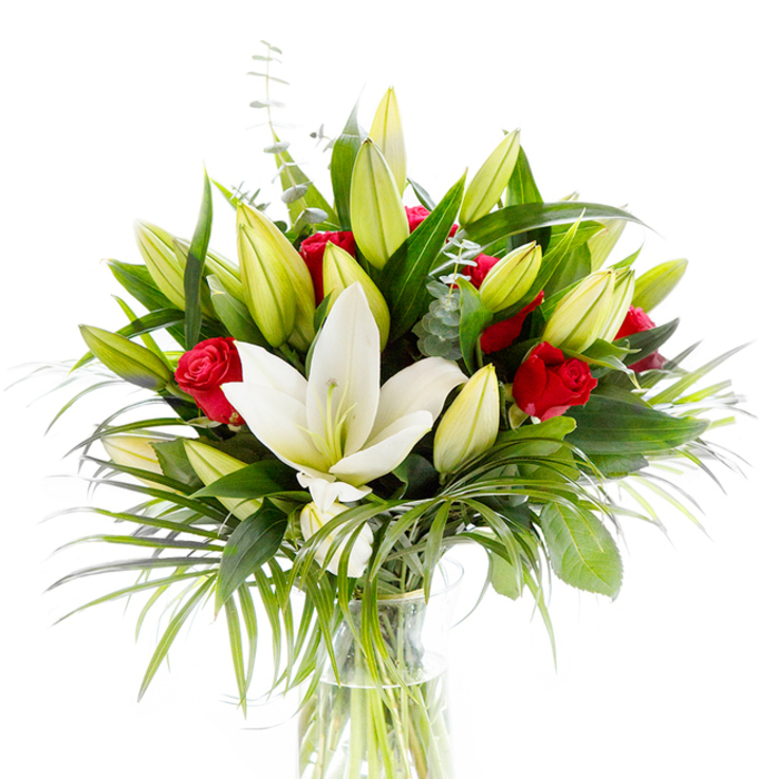 Centered around a single, magnificent white Oriental lily in full bloom, this arrangement balances purity and passion with practiced ease. The lily's immaculate, slightly curving petals frame a subtle yellow stamen and a pale green pistil, the bloom placed forward to catch the eye. Around it sit many lily buds-pale lemon to lime in tone-some still tightly closed and others beginning to unfurl, creating a layered rhythm of expectation. Among these lighter forms, several small, velvety red roses and rosebuds add measured flashes of deep colour; their petals remain mostly curled, lending a demure, classic romance against the lily's brighter statement. The bouquet's foliage is abundant yet refined: large, dark green leaves (likely from the lilies) provide weight and structure; arching palm-like fronds in a brighter emerald hue trail outward to introduce movement; and slim stems of silvery eucalyptus with their characteristic round leaves offer a softer, cooler texture. The whole is composed in a clear cylindrical glass vase, the pale green stems visible beneath fresh water, which gives the arrangement an honest, botanical clarity. Set against a clean white backdrop, the composition's contrasts-soft white, vivid red, and layered greens-are crisp and accessible. The overall impression is one of considered elegance and warmth, the kind of bouquet chosen for anniversaries, romantic gifts, or refined centrepieces. Locally, this style is often requested by clients across Mill Hill and nearby Hale Lane who want a timeless arrangement that reads beautifully in both modern and traditional homes.