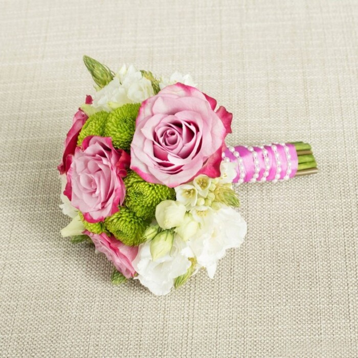 A delicately composed, small bouquet is laid horizontally across a light beige, textured fabric that resembles natural linen or soft burlap, its neutral tone offering a calm stage for the vibrant flowers. Dominating the centre of the design are three open pink roses, two larger and one slightly smaller, each showcasing layered petals that swirl inward to tightly furled hearts. Their colouring moves gently from a pale, romantic blush at the centre to deeper, more saturated pink along the outer edges, with hints of magenta tracing the ruffled petal tips and creating a luxurious ombre effect. Nestled between and around these roses are bright lime-green button chrysanthemums, their spherical, pom-pom forms made up of dense, tiny florets that give a cheerful, contemporary energy to the bouquet. Interspersed among the main blooms are clusters of white and cream filler flowers, likely lisianthus, freesia, or small waxflower, with soft, multi-petaled blossoms and occasional star-shaped flowers sporting tiny yellow centres that add delicate detail and a sense of movement. On the right, the bouquet's hand-tied stems are tightly wrapped in a band of vivid fuchsia satin ribbon, smooth and glossy, providing a striking contrast to the natural textures of both the flowers and the fabric beneath. Over this ribbon, a fine strand of white faux pearls is elegantly spiralled in diagonal lines, reminiscent of a dainty bracelet or bridal accessory, while the neatly trimmed green stems extend just beyond the wrap. The lighting is gentle and even, highlighting the freshness and colour harmony of pink, green, and white. The overall impression is of a chic, intimate bouquet ideal for a Mill Hill wedding, an engagement celebration, or a special event hosted in one of the area's leafy venues, reflecting the refined style often requested from local florists.