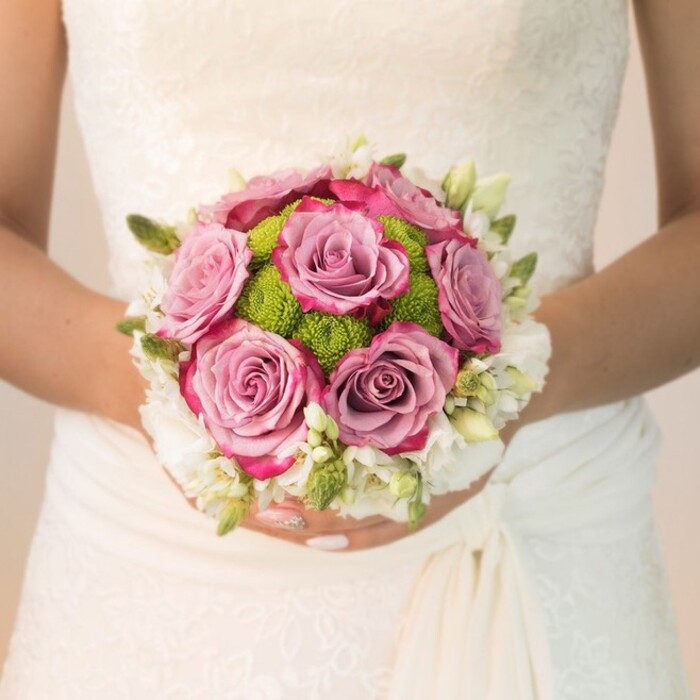 This evocative close-up captures a bride in Mill Hill, her hands delicately encircling a compact round bouquet against the backdrop of an ivory lace dress. The floral gown's intricate overlay hints at local heritage and timeless taste, echoing the refined charm of Mill Hill's tree-lined avenues. At the bouquet's centre, several large roses bloom in a spectrum from dusty lavender through mauve to vivid fuchsia, their ruffled petals almost velvet-like, drawing the viewer's eye and heart. Button chrysanthemums in electric lime-green are clustered throughout, lending texture and an exuberant pop of colour-a touch that reflects the lively greenery found in local gardens. These central flowers are wrapped in a frothy embrace of petite white blossoms, perhaps fragrant freesia or spray roses, with fresh green buds skillfully woven in, their presence adding movement and natural detail. Illuminated by soft, even daylight, the flowers' delicate forms and the bride's well-manicured pink nails, accented by a shimmering detail, radiate a sense of poised celebration. Crafted with the expert care that comes from a deep knowledge of local couples' wishes, this bouquet is ideally suited for wedding inspiration, planning guides, and articles about florists serving the Mill Hill area.