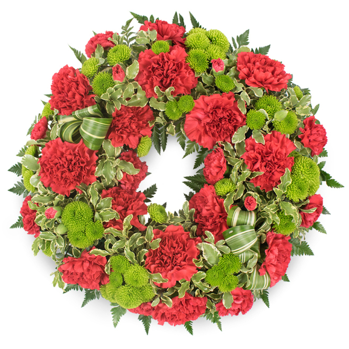 Photographed head-on against a plain white background, this circular funeral wreath appears lush and complete, its form defined by a continuous flow of flowers and greenery with no bare gaps. The colour palette is strong yet soothing: deep red from the carnations and bright, lime green from the button chrysanthemums work together to create a striking but respectful tribute. The carnations are plentiful and tightly arranged, their petals ruffled and layered in soft waves that give each bloom a velvety, almost cloud-like presence. Some flowers have fully opened into wide, rounded circles, while a few are slightly cupped, introducing variety and a sense of natural growth. Grouped between these red blooms are clusters of lime green button mums, their perfectly rounded heads made of tiny, packed petals that catch the light and bring a lively freshness to the design. Beneath and around the flowers lies an intricate underlay of foliage. Dark, feathery fern fronds give the wreath a rich, wooded depth that might call to mind the leafy surroundings of Mill Hill and the green spaces stretching towards the Ridgeway. Interspersed among the ferns, variegated pittosporum leaves with creamy, irregular edges brighten the composition, adding a gentle, patterned contrast. Woven discreetly through the greenery is a ribbon in striped shades of green, folded into soft loops that echo the circular motion of the wreath itself. The whole piece feels evenly balanced, with red and green repeated consistently around the ring, symbolising continuity and the enduring nature of memory. This style of wreath would be a fitting presence at funerals, cremations, and memorial gatherings in Mill Hill, offering a composed, heartfelt expression of sympathy.