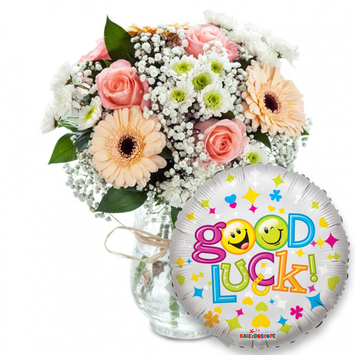 Set against a pristine white backdrop, this cheerful 'Good Luck' arrangement combines garden-fresh blooms and a playful foil balloon to create an immediate sense of celebration and encouragement. The bouquet, seated slightly left of centre in a clear glass vase tied with rustic twine, is crafted from pale peach and soft pink roses whose layered petals suggest a tender, floral perfume. Complementing them are peachy-orange Gerbera daisies with rich dark centres that add graphic warmth, while clouds of tiny white baby's breath (Gypsophila) and tight clusters of white-and-lime chrysanthemum florets provide delicate texture and bright contrast. Deep green foliage peeks through like punctuation, grounding the pastel palette and lending dimension when seen from the front or the side.
Prominently placed to the right and overlapping the vase, a shiny silver foil balloon catches the light and repeats the optimistic message: Good Luck! The balloon's metallic surface is decorated with colourful hearts, stars and confetti shapes in blues, pinks, yellows and greens; the word Good features two sunny yellow smiley faces in place of the 'o's-one cheekily winking, the other sticking its tongue out-while the rest of the letters appear in lively, contrasting hues. The small KALEIDOSCOPE logo is visible at the bottom rim of the balloon. As an arrangement prepared by Flowers Mill Hill, I imagine it being chosen for farewells, exam-day support or new beginnings and delivered with care to addresses across Mill Hill Broadway, Hale Lane and the Village, carrying both colour and thoughtful intent.
