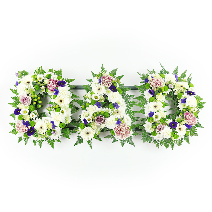 In this photograph, a touching floral tribute spells out the word DAD in three generously sized capital letters, carefully aligned in a horizontal row. Each letter is made entirely from fresh flowers and foliage, with no visible mechanics, giving the impression that the word has been sculpted from petals and leaves. The base of every letter is filled with pure white blossoms: cushion-style chrysanthemums, white gerbera daisies, and similarly petalled flowers that together build a substantial, pillow-like surface. Their petals, some broad and flat, others more tightly packed and feathery, combine to create both softness and definition so the letters remain clearly legible. Surrounding and defining the shapes is a rich collar of greenery-feathery fern fronds and rounded green leaves-which outline each D and the central A, forming a natural frame against the stark white background. Scattered throughout the letters, accent flowers in shades of deep purple and violet punctuate the calm, white field, adding pockets of depth and suggesting quiet strength and dignity. Alongside these deeper tones, clusters of lavender, mauve, and dusky pink roses and carnations bring a gentle warmth, like small echoes of family stories and memories. The lighting is clear and even, minimising shadow and allowing every colour to appear true, much like the kind of light you might find in a peaceful chapel or funeral home in Mill Hill. This floral lettering feels personal and intimate, the kind of bespoke tribute ordered from a trusted local florist such as Flowers Mill Hill to honour a father's life at a funeral, cremation, or memorial gathering. It captures both the formality of farewell and the tenderness of a family's love in one carefully balanced design.