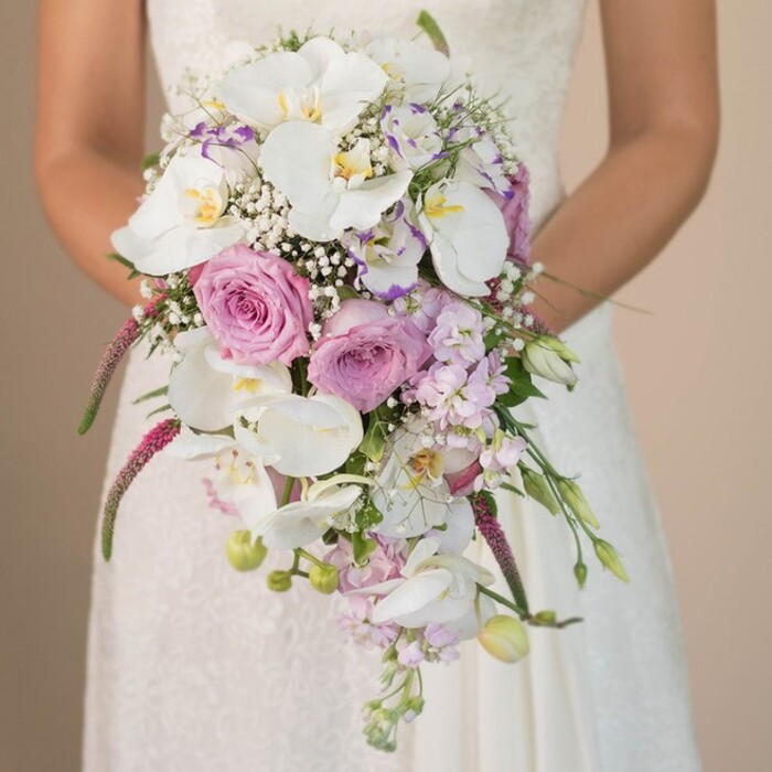 This evocative portrait captures a Mill Hill bride holding a grand, waterfall-style bridal bouquet-an arrangement brimming with colour, texture, and the subtle perfume of classic English flowers. The mid-torso composition invites attention to the details: delicate white and ivory lace on her gown, and the bouquet's luxurious embrace. The design showcases several white Phalaenopsis orchids, their expansive petals and golden hearts gleaming with understated elegance. Surrounding them, layers of lavender and blush pink roses unfold in varying stages, some just budding, others offering their full, velvety allure, reminiscent of gardens bordering Arrandene Open Space. Wisps of white gypsophila interlace the blooms, adding a dreamy, cloud-like fill, while lavender and soft purple blossoms lend a gentle complexity. Trailing fuchsia-mauve amaranthus introduces texture, movement, and artistry to the composition-its bold, tactile presence contrasting beautifully with the surrounding pastels. Fresh green foliage emerges here and there, suggesting both vitality and a link to Mill Hill's leafy charm. The muted background, perhaps echoing the soft palette of NW7's garden squares, allows the bouquet to stand as the undisputed focal point. This creation, a reflection of Flowers Mill Hill's dedication to local brides, celebrates love and hope through its every bloom-an arrangement fit for the most memorable of Mill Hill wedding ceremonies.