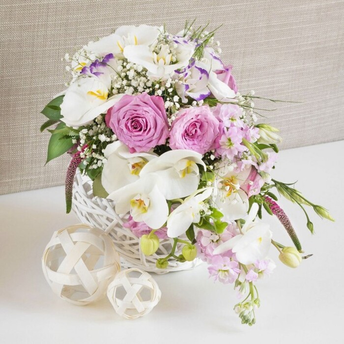 An exquisitely detailed floral arrangement, perfect for a wedding or anniversary celebration in Mill Hill, brims from a rounded white wicker basket that sits on a bright white tabletop. At the centre, lush pink roses open in flawless spirals, each petal unfolding like a soft ribbon, creating a focal point that immediately draws attention. Surrounding them, graceful white Phalaenopsis orchids extend outward, their broad, satiny petals and golden-yellow throats giving the design a luxurious, almost sculpted presence. Intermingled with these statement blooms are ruffled white Lisianthus, some edged with a faint lavender hue, lending subtle variations of tone and a gentle, romantic texture. Stems of pale pink stock emerge and then relax into a mild cascade over the basket's edge, mimicking the natural drape of flowers across a beautifully set table on a quiet lane near Mill Hill East. Delicate sprigs of baby's breath wind through every section of the arrangement, scattering tiny white blossoms like a veil of fine lace between the fuller flowers. Fresh green foliage peeks out here and there, some leaves slender and arching, others more rounded and compact, bringing balance and a lively contrast to the soft colour palette. The white wicker basket, intricately woven and slightly domed, anchors the arrangement with a hand-crafted charm. To one side, two decorative spheres made from pale, interlaced wood sit on the same white surface, their open, airy construction echoing the weave of the basket and adding another layer of texture. Behind, a light beige, gently textured fabric backdrop remains unobtrusive, allowing the pinks, whites, and fresh greens to shine. The overall scene feels calm, polished, and joyfully romantic, ideal for illustrating wedding table arrangements or special occasion florals in Mill Hill.