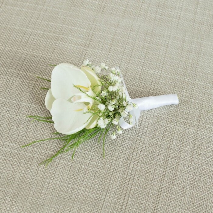Resting on a softly woven, oatmeal-beige fabric that looks like natural linen, this elegant white orchid boutonniere appears calm and perfectly composed, much like a quiet afternoon in Mill Hill's leafy lanes. At the centre lies a single Phalaenopsis orchid bloom, its petals broad, smooth, and slightly luminous, curving outwards in a gentle, sculptural form. The pure white petals are unblemished, with a faint sheen that catches the light, while the heart of the flower reveals a delicate touch of pale yellow and a hint of cream, adding warmth and a subtle focal point. Encircling the orchid are airy clusters of white baby's breath, each Gypsophila floret minuscule yet crisp, creating a soft, cloud-like halo around the main bloom. The texture of the baby's breath contrasts beautifully with the orchid's velvety surface, lending the arrangement a sense of lightness and romance, as if a fine mist had settled around it. Slender strands of fresh green foliage peek between the tiny white blossoms, the fronds fine and feathery, suggesting a delicate fern or similar leafy detail. These green accents add a lively touch, reminiscent of the greenery around Mill Hill Park or the gardens near the Ridgeway, and help ground the design in nature. The stems are tightly aligned and wrapped with a pristine white satin ribbon, smooth and slightly glossy, wound neatly from base to just below the orchid and finished with a small, discreet bow. The lighting is soft and even, with no harsh shadows, allowing the crisp whites and bright greens to be clearly defined against the neutral background. Crafted with the attention to detail you'd expect from an experienced Mill Hill florist, this boutonniere feels ideal for a groom, groomsman, or prom guest seeking a refined, timeless accessory for a formal celebration in north London.
