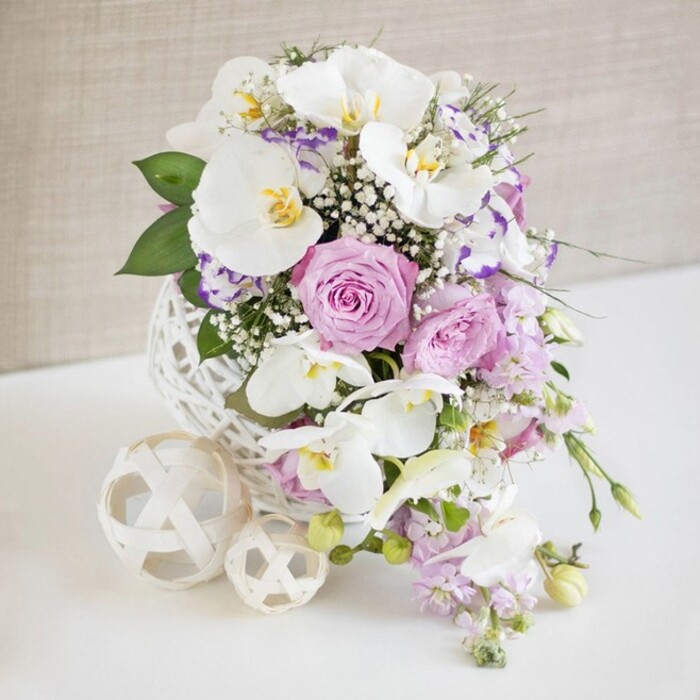 An exquisitely arranged floral display is photographed against a gentle, neutral backdrop, evoking the calm elegance of a Mill Hill celebration at dusk. The arrangement stands in a round, white decorative vase, crafted from slender strands that criss-cross to form an open, woven sphere reminiscent of delicate rattan or fine wicker art. This sculptural vessel, placed on a pristine white surface, becomes part of the design, its texture mirrored by two nearby cream-coloured woven balls that add balance and a touch of modern minimalism. From the vase, a lush bouquet of orchids and roses rises in a rounded, slightly overflowing shape. Several large white Phalaenopsis orchids anchor the composition; their petals are broad, smooth, and gently curved, with soft yellow and golden accents glowing at their centres, giving the flowers a refined, almost luminous presence. Encircling these orchids are roses in shades of blush pink and light lavender, some tightly furled, others fully open to display a rich tangle of petals suggestive of romance and celebration. Smaller white and lilac ruffled blooms, reminiscent of Lisianthus with delicately coloured edges, peek between the larger flowers, adding texture and subtle tonal shifts. Threaded throughout are airy sprays of white baby's breath, creating a silvery, cloud-like veil that softens the whole design. Delicate stems of light purple blossoms trail gently downwards, introducing a graceful cascade that breaks the symmetry and suggests natural movement, like flowers drifting towards the edge of a terrace near Mill Hill's green spaces. Rich green foliage frames and supports the pastel blooms, providing depth and freshness. The light is bright yet diffused, with no harsh shadows, so every petal, weave, and leaf is clearly defined, perfect for representing wedding table arrangements or luxurious events in Mill Hill.