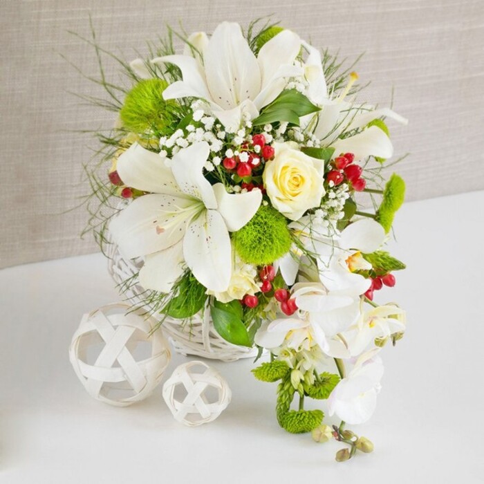 This image shows a meticulously crafted table arrangement composed primarily of white blooms and lush greenery, arranged in a round white woven basket that sits on a polished white surface. The scene is framed by a softly textured beige background, which recedes gently so that the flowers command full attention, as they might on a top table at a Mill Hill wedding venue. Dominating the upper section of the bouquet are several striking white lilies, their large, open petals slightly ruffled at the edges and delicately freckled near the centre. Their stamens, tipped with golden anthers, arch outward and give the impression of a light, perfumed presence. Near the upper middle sits a single, creamy pale-yellow rose, its petals cupped and layered in a classic spiral, offering a warm, romantic counterpoint to the cooler white tones around it. Interwoven with these focal flowers is a mix of foliage: fine, feathery greens that stretch outward in soft lines, and broader, darker leaves that anchor the base. Airy sprigs of white baby's breath appear throughout, their tiny blossoms creating a gentle haze that softens transitions between the larger blooms. From the right, a graceful cascade of white Phalaenopsis orchids flows downwards, their sculptural petals and elongated stems forming a waterfall effect that lengthens the arrangement and adds a sense of movement. Bright green, spherical pom-pom flowers nestle within the greenery, their unusual texture suggesting moss-like cushions and adding visual intrigue. Threaded among the foliage and blossoms are clusters of small, rich red berries, which catch the light and introduce a festive vibrancy without overwhelming the design. To the left of the basket, two white decorative balls made from interwoven strands rest on the table, echoing the wicker and adding a minimalist, contemporary note. The overall impression is of refined luxury and timeless elegance, well suited to formal celebrations and special events across Mill Hill and nearby Hendon.