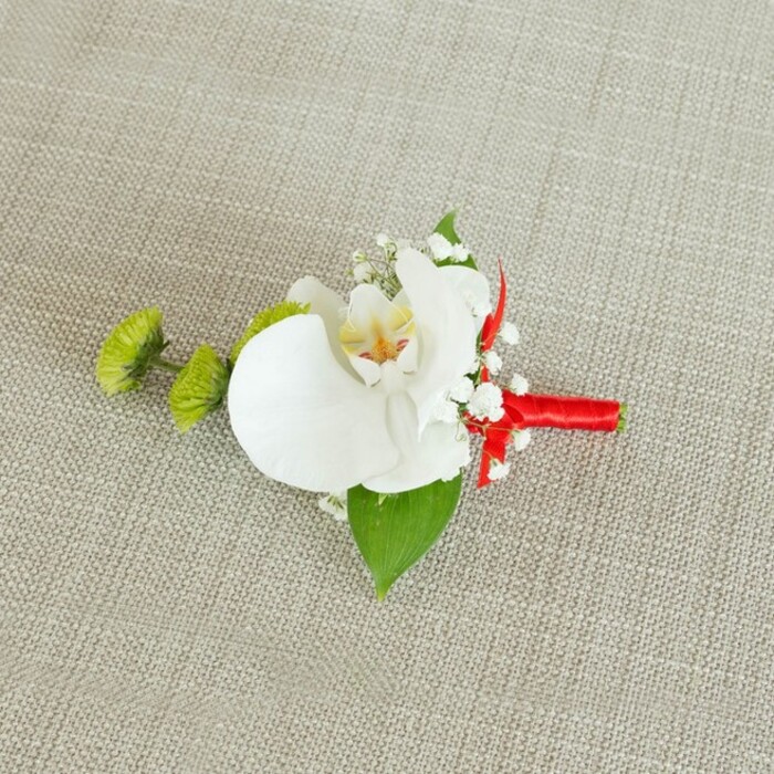 An elegant wedding boutonniere is presented against a softly textured, light beige linen-style fabric, its understated background allowing every detail of the flowers to stand out with clarity and grace. At the centre, a large white Phalaenopsis orchid opens in full bloom, its petals broad and gently overlapping, like folded silk illuminated by soft daylight. Inside the flower, the throat glows with warm yellow tones that gradually deepen into delicate streaks of reddish-orange and fine brown speckling, adding a painterly touch to the otherwise pristine white. Surrounding this central orchid, airy sprigs of baby's breath scatter tiny white blossoms, creating a halo of cloud-like texture that softens the edges of the design. On the left side, two compact, round lime-green chrysanthemum buds, with their tightly coiled, spiky petals, offer a refreshing pop of colour and a distinctly modern, textural contrast. Beneath the blooms, a single broad, deep green leaf curves like a subtle backing board, grounding the composition and echoing the verdant foliage you might notice in the landscaped corners of Mill Hill and along its quiet residential streets. Closer to the stems, another smaller, darker green leaf peeks through, adding depth and variation in tone. The stems are neatly bound in a vivid crimson red satin ribbon, wrapped with practiced precision and ending in a small knot or discreet bow that introduces a bold, celebratory note of colour. This juxtaposition of white, green, and red creates a harmonious, eye-catching accent that would look perfectly at home on a tailored lapel at a Mill Hill wedding or formal event. Crafted with the meticulous care you'd expect from an experienced local florist, the boutonniere balances tradition and contemporary style, ideal for capturing the significance of vows, celebrations, and milestone occasions.