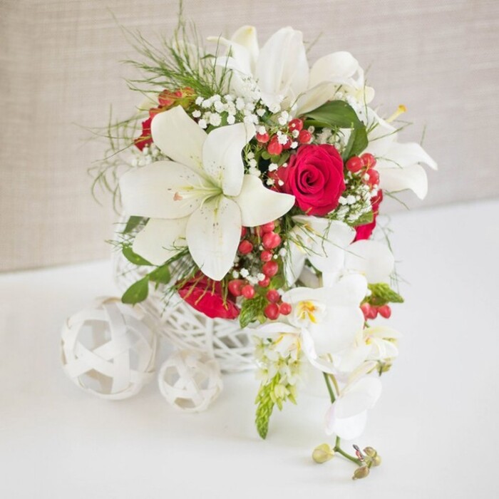 A beautifully composed floral centerpiece, reminiscent of a bridal arrangement ready to grace a Mill Hill reception table, is carefully staged against a soft, light beige backdrop that allows the colours to glow. The flowers are nestled in a low, round basket-style container made from intricately woven white strands, its open, airy pattern adding interest without visual weight, much like delicate décor chosen from a favourite North London artisan shop. Rising from this base, white oriental lilies take centre stage, their petals wide and graceful, slightly recurved at the tips, with prominent stamens that carry a dusting of warm pollen. These lilies feel both classic and modern, forming a luminous frame around the deeper tones. Between them sit velvety red roses, rich in colour and tightly layered, creating pockets of romantic warmth that would be perfect for anniversaries or special dinners at homes near Mill Hill Park. Fine white gypsophila, or baby's breath, floats through the arrangement, softening edges and creating a gentle, cloud-like veil among the bolder blooms. Wispy fronds of fresh green foliage curl and weave through the bouquet, adding brightness and giving the impression of freshly cut stems, just arranged by experienced local florists at Flowers Mill Hill. Nestled here and there, glossy red hypericum berries bring playful, festive dots of colour, harmonising with the roses and enhancing the seasonal feel without shifting the palette away from its elegant simplicity. To the right of the main cluster, a stem of white phalaenopsis orchids cascades downward, each bloom pristine and smooth, marked at the centre with touches of yellow and pink, while small buds at the tip hint at flowers yet to open. On the white surface to the left, three decorative spheres, also made from white woven material, echo the pattern of the container and complete the thoughtfully styled scene. The overall effect is one of refined luxury and intimate celebration, ideal for wedding breakfasts, engagement parties, or sophisticated gatherings in Mill Hill.