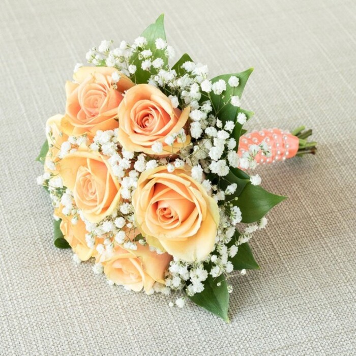 Laid gently across a light, tactile linen reminiscent of intimate Mill Hill gatherings, this bridal bouquet combines classic romance with local charm. About seven radiant peach roses, unfolding their layers from tight centers to luxuriously soft, ruffled edges, are thoughtfully arranged in a compact, rounded form. Interwoven throughout are myriad delicate tufts of baby's breath-minute white florets lending a gossamer, floating effect. These ethereal details create a striking contrast against the vibrant, warm tones of the roses, while rich, deep green leaves offer structural depth, calling to mind Mill Hill's ancient hedgerows and greenways. The handle, destined to nestle within a bride's or bridesmaid's grasp, is swathed in a smooth, coral-peach ribbon complimented by a dainty spiral of glistening white pearls, infusing the bouquet with an accent of ceremonial luxury found at Pine Road receptions or events along Mill Hill Broadway. The exposed, freshly cut stems beneath reinforce the bouquet's authenticity and the promise of freshness from local florists. Illuminated by soft, even light, every petal, pearl, and textural fold is revealed in vibrant detail, inspiring feelings of tenderness and grace suited to wedding celebrations or anniversary moments within the heart of Mill Hill.