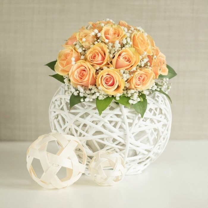 A stylish and serene floral composition presents a softly rounded bouquet of peach and apricot-toned roses, their petals tightly swirled and layered to form plush, velvety blooms that sit closely together in a harmonious dome. The centers of the roses are a richer, sun-warmed orange, gradually dissolving into paler peach and cream at the edges, giving the arrangement a subtle ombré effect that feels both romantic and contemporary, ideal for a chic gathering in a Mill Hill garden flat or a small celebration near Arrandene Open Space. Interspersed generously between the roses are small, cloud-like tufts of white baby's breath, each cluster a constellation of tiny blooms that lend airiness and movement, as if the flowers were gently breathing. A few glimpses of deep green foliage peek out near the base of the bouquet, grounding the pastel tones and adding quiet depth. The flowers are nestled in a distinctive spherical vase crafted from white woven strands that crisscross in an open, lattice pattern, rather like refined wicker or rattan. This sculptural container, with its see-through structure and curved silhouette, brings a touch of modern design to the otherwise classic combination of roses and gypsophila. Resting on the crisp white surface in front of the vase are two smaller decorative spheres made from the same white material: one larger orb, neatly patterned and reminiscent of a simplified football, and a smaller, more irregular ball that introduces a playful, artistic note. The backdrop is a soft, pale beige or greige wall with a faint vertical texture, keeping the setting calm and uncluttered so the warm roses and intricate white weaves remain the clear focal point. Gentle, diffuse daylight illuminates the scene, suggesting a carefully curated interior in NW7, ready for guests arriving from Mill Hill Broadway or from nearby Barnet for a relaxed yet polished celebration.
