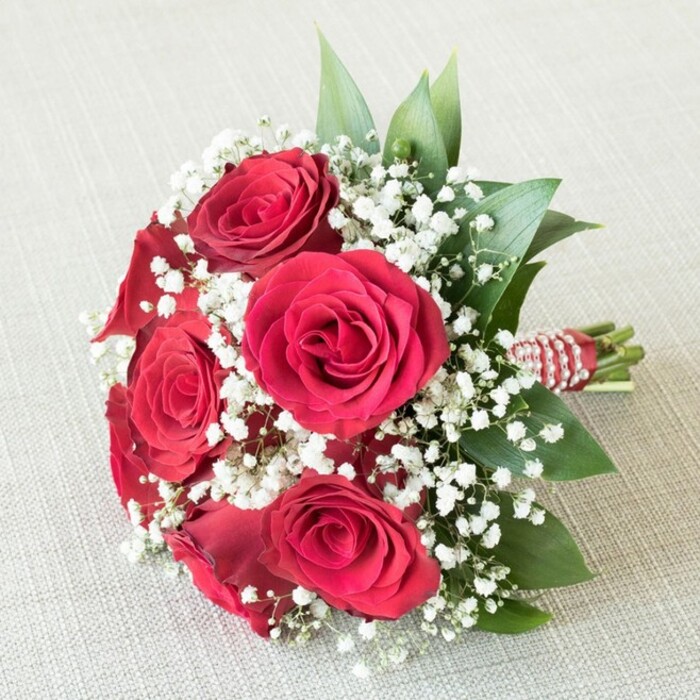 Set against a soft, neutral-toned fabric that calls to mind the peaceful afternoons in local Mill Hill gardens, this exquisite bridesmaid bouquet from Flowers Mill Hill captures the essence of timeless romance. The centerpiece showcases six premium, deeply crimson red roses, their velvety petals tightly furled and unfolding elegantly, offering a rich tapestry of colour and texture. Ample clusters of pristine white baby's breath (gypsophila) are woven throughout, their airy blooms forming a gentle mist that softens the intensity of the roses. Chic, glossy foliage peeks through and surrounds the flowers, imbuing the bouquet with vibrant life and echoing the verdant charm of Mill Hill's fields and leafy avenues. Skillful binding at the bouquet's base features a vivid red and white striped ribbon, tastefully adorned with a delicate twist of pearly white beads, lending the arrangement both sophistication and a conversational, local flair. Subtle, even lighting accentuates each component, allowing the rich reds, bright whites, and fresh greens to glow in harmonious balance. This visually striking bouquet offers an ideal expression of love, gratitude, or celebration within Mill Hill, thoughtfully crafted for memorable weddings and joyful gatherings.