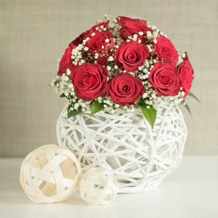 A beautifully composed floral scene fills the frame, evoking the quiet anticipation of a wedding breakfast table near Mill Hill East. Dominating the centre is a rounded bouquet of deep red roses, a tightly arranged cluster of roughly a dozen to fifteen blooms. Each rose is at its best, fully open with spiralling petals that curl gently outward, creating a velvety, layered surface that looks almost like rich fabric. Nestled between these crimson blossoms are fine sprays of white baby's breath, or gypsophila, their tiny, clustered flowers forming a soft, misty backdrop that lightens the overall effect and adds a lacy delicacy. Every so often, small glimpses of green foliage emerge from the mass of petals, grounding the design in its natural origin and adding a gentle, contrasting depth. The bouquet is held aloft by a distinctive white vase, round and slightly bulbous, constructed from interlaced white strands that cross and loop to create an open, woven lattice. The structure resembles painted wicker or rattan shaped into a modern, sculptural container, its bright white surface harmonising beautifully with the baby's breath and making the red roses appear even more vivid. Resting on the smooth, pale tabletop beside the vase are two decorative woven spheres in soft cream and beige hues, one larger and one smaller; their airy, open weave echoes the design of the vase and adds a calm rhythm to the scene. The background is a neutral, gently textured wall that fades out of focus, much like the soft, distant view you might glimpse from a Mill Hill garden room. The overall feeling is one of relaxed sophistication and romance, suitable as a wedding table arrangement, anniversary centrepiece, or elegant decor for celebrations in and around Mill Hill.
