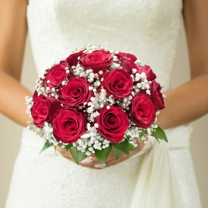 A moment of Mill Hill bridal anticipation is exquisitely captured as a bride holds a bouquet that blends tradition with local artistry. At the heart of the arrangement, an opulent array of deep red roses stands proudly, each bloom meticulously layered and glowing with a rich, velveteen texture. Pockets of baby's breath-each tiny blossom a puff of snowy white-give the bouquet a soft, cloudlike allure. Underneath, the subtle emergence of fresh green leaves adds a lively touch, mirroring the gardens and leafy lanes of Mill Hill. The bride's perfectly groomed hands enclose the stems, her manicure immaculate, while the floral arrangement rests gently against the intricate, lace-embellished bodice of her white gown. A soft sash falls gracefully along her side, its movement suggested by gentle light filtering through a blurred, creamy backdrop. Every element-the crispness of the flower petals, the sweep of wedding fabric, and the natural accents-converges into a scene of intimate joy and artistry. This image is perfect for illustrating contemporary bridal elegance, local Mill Hill traditions, or the enduring appeal of classic wedding bouquets.