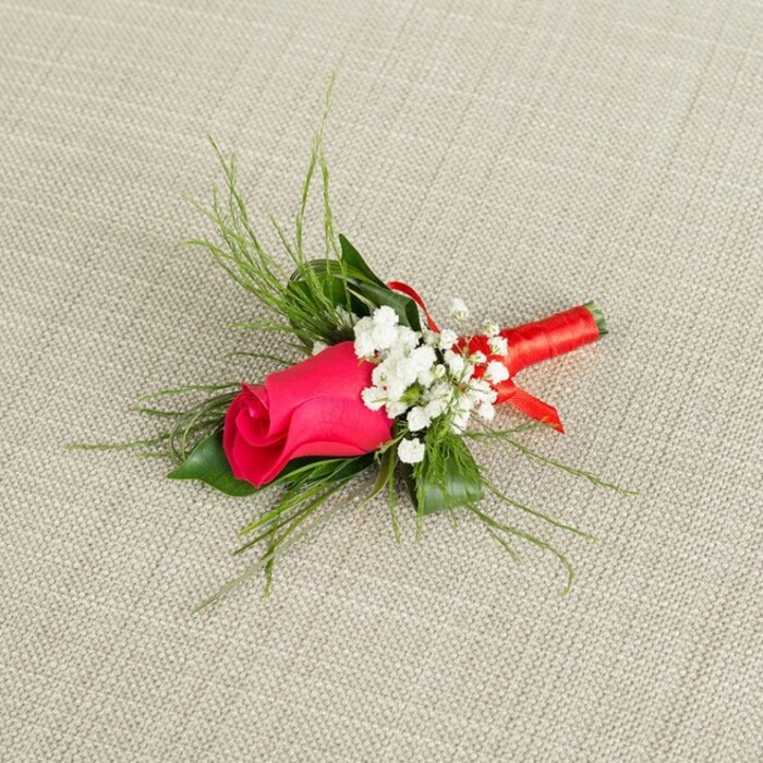 On a pale, natural-looking fabric background, similar to finely woven linen in a warm beige tone, a single-stem boutonnière is arranged with the precision and care you would expect from an experienced Mill Hill florist. The focal point is a tightly closed rosebud in a vivid fuchsia or deep magenta shade, its petals smooth, layered, and gently curling at the edges, suggesting freshness and the anticipation of opening later in the evening. Framing this central rose, clusters of tiny white baby's breath blossoms gather like a soft veil, each miniature flower lifting the design with airy brightness and offering a gentle contrast to the rose's saturated colour. Around and behind the blooms, a combination of greenery gives depth and movement: slim, grass-like fronds and feathery, bright green sprigs arch outward, while a couple of broader, darker green leaves cradle the base, echoing the lush foliage of Mill Hill Park after spring rain. The stems are pulled together into a compact bundle and wrapped smoothly in a rich red satin ribbon, the glossy fabric hugging the stems in a firm, elegant band that glints subtly where the light catches it. The boutonnière lies at a slight diagonal across the image, mirroring how it would naturally sit on a lapel at a wedding, prom, or special celebration dinner on Mill Hill Broadway. The lighting is clean and bright, with no harsh shadows, allowing you to sense the velvety touch of the rose, the crispness of the baby's breath, and the cool, silky feel of the ribbon, all combining to convey romance, refinement, and a timeless sense of occasion perfect for formal wear.