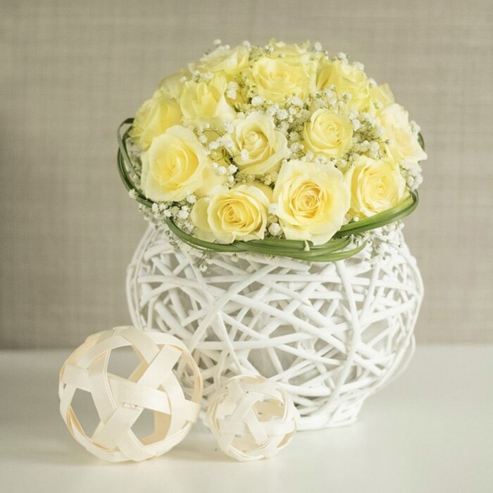 A beautifully balanced floral display rests on a smooth, reflective white tabletop, its focal point a perfectly rounded mound of soft yellow roses. The blooms range from buttery cream to gentle primrose, each rose carefully opened so the petals form layered spirals, creating a rich yet orderly surface of colour and texture. Interspersed throughout the dome are clusters of delicate white baby's breath, their countless tiny blossoms giving the arrangement an airy, cloud-like softness, as though a faint mist has settled among the petals. Encircling the base of the bouquet is a slender band of vivid green foliage, perhaps neatly arranged flat leaves, that acts as a natural border, adding definition and a refreshing hint of garden greenery. The flowers are supported by a distinctive white woven vase, its shape gently curving in at the centre like an elegant urn. Slim strands intertwine to form an open, basket-style pattern, allowing light to pass through and casting fine, crisscrossed shadows on the table beneath. Nearby, two spherical decorative accents crafted from pale, rattan-like strips continue the open-weave motif; the larger ball sits slightly forward, with the smaller tucked beside it, echoing the vase's texture and bringing harmony to the composition. Behind this centerpiece, a softly blurred, neutral beige background ensures the eye remains focused on the floral variation, the woven materials, and the interplay of white, cream, and green. Created with the care of experienced North London florists, this arrangement would suit a wedding reception in Mill Hill, a stylish celebration near The Ridgeway, or a serene home setting, radiating gentle joy and contemporary elegance.