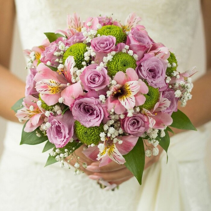 A striking, hand-tied bridal bouquet is gently clasped by a bride, who stands in a flowing white dress against the soft background of embroidered lace-a familiar scene from cherished Mill Hill weddings. This bouquet marries elegance and modernity, featuring a harmonious medley of blush pink and lavender roses at its centre, their petals dewy and fragrant from the hands of local florists near Mill Hill Broadway. Interlaced between the roses, lime green button chrysanthemums offer spirited pops of colour and spherical texture, lending a contemporary accent that sets this arrangement apart. Pink alstroemeria lilies, marked with fine maroon streaks and blush centers, are artfully positioned, their elongated petals curving gracefully around clusters of tiny white baby's breath, whose delicate blooms hover throughout the bouquet, imparting a sense of airiness and romance. At the structure's base and edges, glossy dark green leaves peek out, their lushness framing the pastel flowers and adding depth. Illuminated by the gentle daylight of a spring morning in Mill Hill, each element reveals its freshness and natural beauty. This bouquet conveys joy, hope, and artistry-perfect as a centrepiece for a local wedding, a muse for bridal inspiration articles, or a statement example of Flowers Mill Hill's bespoke service for celebrations throughout the area.