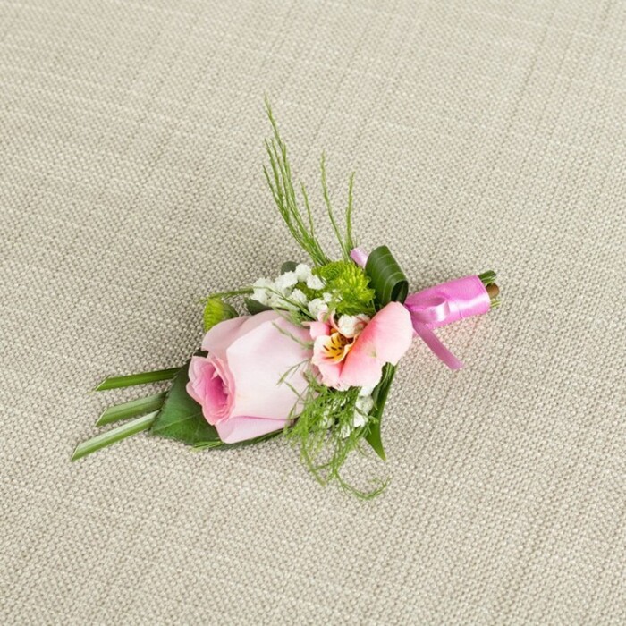 A sophisticated floral boutonniere, assembled by a skilled Mill Hill florist, reclines diagonally on an understated, pale beige fabric-the weave suggesting the quiet refinement of a local gallery space. The arrangement's heart is a soft pink rosebud: its tightly rolled petals a testament to freshness and latent romance. Surrounding the rose delicately, sprays of tiny white Gypsophila bring classic, airy lightness to the design. Miniature, vibrant green spherical mums introduce a pleasing contrast and hint of spring, reminiscent of walks along Arrandene Open Space. A gently ruffled, light blush or peach blossom with dewdrop-yellow accents peeks out-perhaps an alstroemeria or lisianthus-contributing both colour and awhile subtle intricacy. Wispy fern fronds and slender, emerald grass blades arch gracefully upwards, their energy softened by the richness of broader, flatter leaves forming the base, including an expertly folded leaf for added dimensions. At the binding, a lustrous fuchsia satin ribbon is tied into a petite and tidy bow, shimmering in natural light. One broad green leaf is artfully wrapped around the gathered stems, framing the arrangement with a final flourish. Presented against a textured, neutral textile, the boutonniere's layered hues and varied textures stand out. It is a thoughtfully crafted accent for a Mill Hill wedding, school prom, or formal event, ready to be pinned to a lapel as a symbol of romance, elegance, and the lush charm unique to the Mill Hill area.