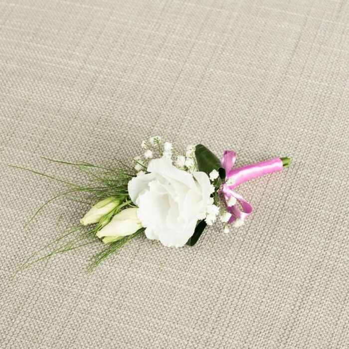 Set against a softly woven, light beige fabric that recalls the understated elegance of table linens in Mill Hill's intimate wedding venues, a refined boutonnière rests at a gentle diagonal, composed with the practiced hand of an experienced florist. The focal bloom is a fully open white lisianthus, its layered, ruffled petals unfolding in delicate spirals that create a lush, rounded form. The petals are creamy at their base, brightening to a crisp white along the edges, and they catch the light in a way that makes the flower appear almost luminous against the neutral background. Just below and slightly to the left, two slender lisianthus buds, tinged with pale green, are nestled closely to the main flower, their tapered forms suggesting blossoms still waiting for their moment-perfect symbolism for a couple just starting their journey, perhaps exchanging vows near Mill Hill Park or overlooking the greenery by Arrandene Open Space. Interspersed throughout the composition are airy tufts of white gypsophila, each tiny flower dotting the arrangement with a star-like delicacy that softens the overall silhouette. Backing the central cluster is a deep green, smooth leaf, its broad shape grounding the design, while a few fine, grass-like strands of foliage extend towards the lower left, adding a sense of movement and a whisper of wildness, as though freshly gathered from a local garden. At the base, the slender stems are neatly bundled and wrapped in a vibrant fuchsia pink satin ribbon, finished with a small, playful bow that adds a shot of modern colour and a subtly romantic flair. The photograph, taken from an overhead angle with gentle, even lighting, highlights every texture-from the satin's sheen to the fabric's subtle weave-conveying a sense of poised calm before the excitement of a wedding or prom in Mill Hill.