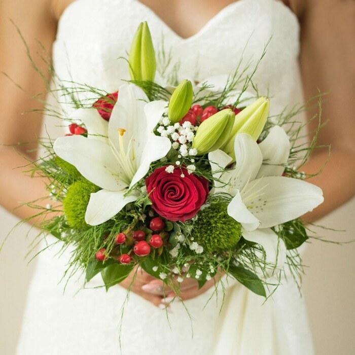 In this detailed composition at a Mill Hill wedding venue, a bride's hands are folded gracefully around a bouquet bursting with traditional and modern touches. The star of the arrangement is the array of pristine oriental lilies, their large white petals unfolding elegantly, some with golden stamens visible, others as pale green buds indicating the promise of days to come. At the bouquet's center rests a deep, velvety red rose-a focal point that radiates passion against the creamy floral backdrop. Wisps of delicate baby's breath (Gypsophila) are scattered throughout, softening the look and creating a dreamlike air, while clusters of button-like green trick dianthus provide playful, mossy texture. Red hypericum berries, small yet vibrant, punctuate the scene with joyful colour-echoing the energy found in Mill Hill's markets and gardens. The design is finished with a rich variety of foliage, from slender asparagus fern fronds to bold leafy greens, creating a full, gathered effect reminiscent of English countryside walks. The image is softly lit, focusing attention on the lush floral details and the quiet anticipation held in the bride's hands. Designed with the Mill Hill community in mind, this bouquet blends classic wedding elegance with a fresh, lively spirit, offering inspiration for couples seeking meaningful floral artistry for their special day.
