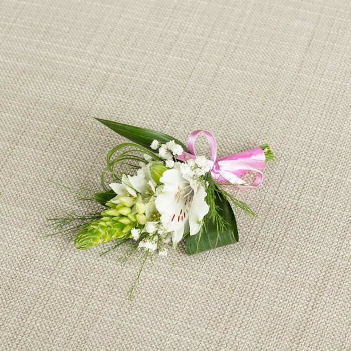 A meticulously crafted boutonniere, likely destined for a wedding or prom in Mill Hill, is displayed on a subtly textured, light beige fabric that resembles finely woven linen, its neutral tone providing a serene, almost gallery-like backdrop. The central focus is a single white alstroemeria blossom, sometimes known as a Peruvian lily, its petals broad and slightly undulating at the edges, catching the soft light in a way that highlights their almost translucent quality. Near the throat of the flower, delicate reddish-brown speckles fan out like careful brushstrokes, adding character and depth to the otherwise pristine white petals, while the stamens sit gently at the centre, hinting at a light, green-fresh fragrance. To the left of this main bloom, a striking, conical spike of bright green buds rises from the arrangement, resembling an Ornithogalum or similar structured flower, its tight, layered segments bringing a bold, modern texture to the piece. Around these focal elements, clusters of baby's breath weave in and out, their minuscule white flowers appearing as a soft mist, lending a dreamy, romantic air. The greenery is thoughtfully layered: strong, dark green, spear-shaped leaves form a sturdy base that cradles the flowers, while slender, grass-like strands arc outward and upward, introducing elegant lines and a sense of gentle movement. At the right-hand edge, the stems are neatly bound with a glossy pink satin ribbon tied into a small, perfectly formed bow, adding a cheerful yet sophisticated note of colour. Captured in even, natural-style lighting, the boutonniere feels freshly made, as though it has just left the workbench of a local Mill Hill florist, ready to be pinned onto a lapel for a ceremony, family celebration, or special photograph on the leafy streets and green spaces nearby.