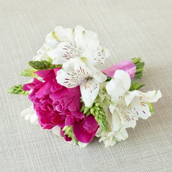 Displayed against a softly woven, light beige background that resembles natural linen, this small floral arrangement appears as if it has just been gently set down between photos at a Mill Hill wedding. The neutral fabric, with its subtle texture and warm undertone, creates a calm canvas for a vivid cluster of blooms tied neatly together. At the centre, a striking fuchsia peony-style flower is in full, generous bloom, its many ruffled petals stacked in rich layers of magenta and pink, giving it a plush, almost cloud-like appearance. Surrounding this central blossom are several pristine white alstroemeria, or Peruvian lilies, their elongated petals slightly reflexed at the tips. Each lily is marked with fine burgundy or maroon streaks and speckles that gather towards the throat, as if brushed on by hand, adding a sense of intricate detail and quiet drama. Intermingled among these focal flowers are slender stems of vivid green buds and spiky florets, offering a fresh, vertical texture that contrasts with the rounded softness of the peony. The stems of the entire cluster are carefully aligned and wrapped in a narrow band of bright pink satin ribbon, its smooth, glossy finish reflecting light and echoing the peony's rich hue. Gentle, even lighting bathes the arrangement, casting soft shadows that define each petal's curve while keeping the overall mood light and romantic. The piece feels like a polished boutonnière or small posy crafted by an experienced local florist serving Mill Hill, perhaps destined for a lapel at a celebration near Mill Hill Park or a family gathering off Bunns Lane, offering a sophisticated yet joyful burst of colour and texture.