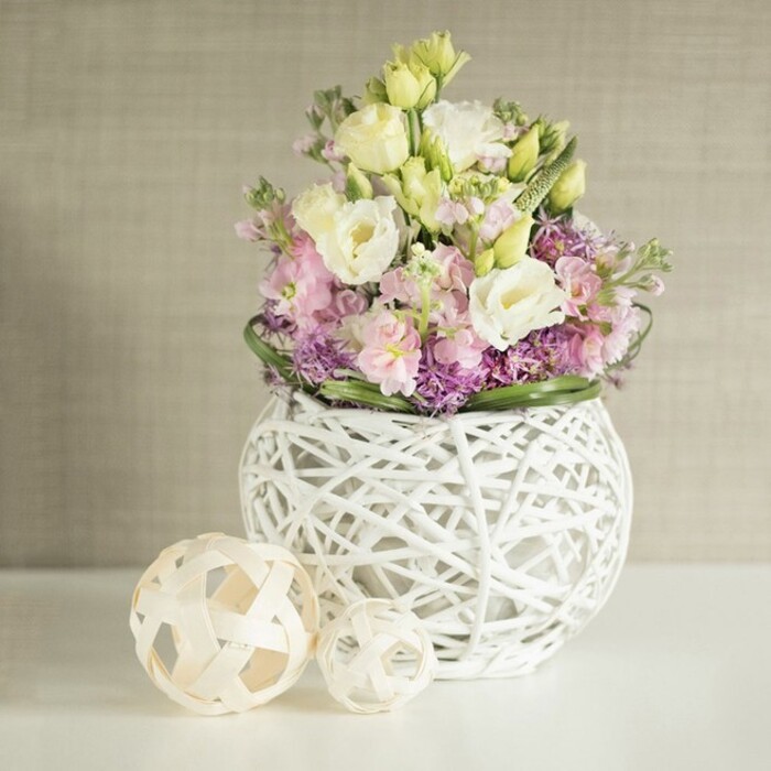 Distinctive in its airy, sculptural beauty, this lavish flower arrangement draws in the spirit of Mill Hill's leafy charm and suburban sophistication. Central to the bouquet are velvety lisianthus blossoms in shades of white and pale yellow, their layered petals radiating subtle light and a sense of refinement. Towering among them, soft pink stock flowers cast poetic silhouettes, bringing a vertical dynamic that suggests growth and optimism-echoes of green spaces around NW7. Deep violet and mauve filler blooms, possibly allium or aster, add plush texture beneath, allowing the arrangement to feel abundant yet never crowded. The verdant edge of lobed green foliage sets a crisp border, drawing the eye back to the centre. Everything is arranged within a spherical openwork vase-its white rattan weave reminiscent of local artisanship found at Mill Hill crafts fairs-sitting atop a gleaming white table. Nearby, two woven decorative spheres further the artisanal mood, completing this scene of creative unity. The backdrop is a soft greige wall with subtle vertical threading, offering an understated canvas that amplifies the bouquet's hues. Perfect for refined gatherings, wedding celebrations, or as an uplifting centrepiece delivered locally by Flowers Mill Hill, this tableau is a testament to the neighbourhood's cultivated taste and the florist's skilled hand.