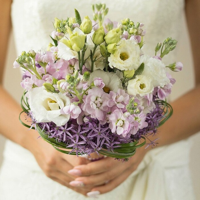 Every petal, every stem in this close-up bouquet portrait reflects the refined grace of Mill Hill weddings. The arrangement radiates with creamy white Lisianthus, their ruffled petals and light greenish-yellow centers shining softly, capturing the morning calm found on the edges of Mill Hill Park. Generous tufts of pastel pink and lilac stock interlace between the Lisianthus, contributing romantic texture and a gentle, frothy depth reminiscent of the local flora during spring. Unopened Lisianthus buds, fresh and green, peek through, bringing an element of new beginnings. At the bouquet's base, a collar of deep purple, spiky Allium forms a mesmerizing and structured foundation, providing a crisp architectural line that aligns with the curated beauty of Mill Hill's landscaped spaces. Loops of sleek, verdant bear grass are woven around the midpoint, adding modern structure and a nod to the green expanses found along Bittacy Hill. The bride's fingers, neatly manicured in soft blush, cradle the bouquet's wrap, while the background fades into blurred ivory-perhaps lace or softly textured gown fabric. Crafted by a local floral designer and thoughtfully styled, this bouquet radiates celebration and hope, an exquisite fit for ceremonies and celebrations in NW7's welcoming surroundings.