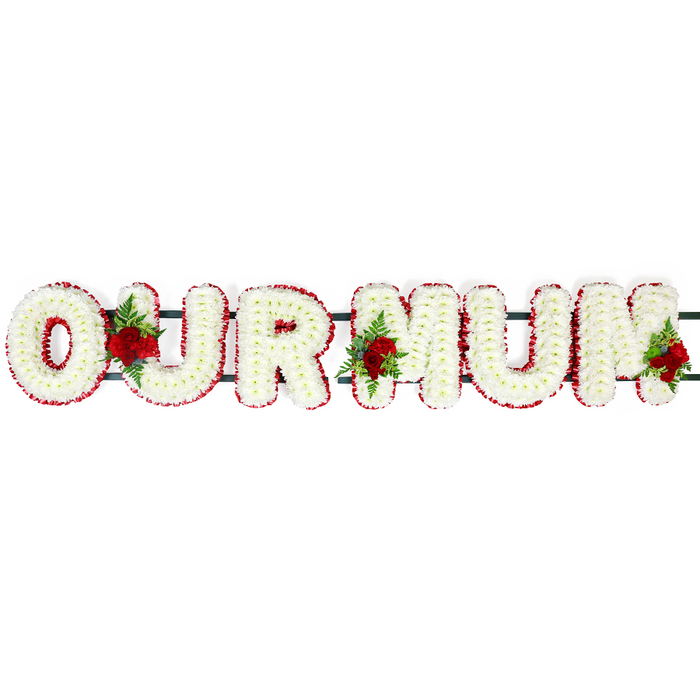 This image presents a traditional OUR MUM funeral flower arrangement, the kind of tribute you might see at a service in Mill Hill, arranged with precision and care. The letters are solid block capitals, positioned in a single row on a hidden support that appears only as a dark green bar along their base. The faces of the letters are completely covered in small white chrysanthemum florets, each bloom tightly nestled against the next. Their layered petals fan out to create a plush, almost quilted appearance, with tiny shadows in the crevices between blooms lending soft dimension. Around the edge of every letter runs a narrow yet prominent border of deep red flowers, likely spray carnations or similar small-headed blooms. This border forms a crisp halo around the white centre, emphasising the shape of each character so the words OUR MUM can be easily read even from a distance. Three decorative rose sprays punctuate the tribute, adding both colour and emotional weight. Clusters of deep red roses, their petals rich and velvety, are positioned between the O and U, the U and first M, and at the end of the final M. These roses sit in a bed of lush green foliage and delicate fern, which spills slightly over the white chrysanthemums, softening the edges and bringing a garden-like feel. The combination of white, red, and green evokes purity, devotion, and remembrance, capturing the sentiment of a final goodbye to a cherished mother. You might imagine this arrangement placed lovingly beside the coffin in a church near Mill Hill Broadway, or resting at a graveside in the quiet grounds around Mill Hill, where the still air and birdsong heighten the sense of reflection and love.