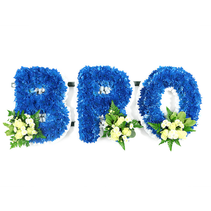This studio image captures a heartfelt memorial arrangement spelling BRO in three separate, freestanding letters, each lavishly formed from compact royal blue chrysanthemums. Viewed at eye level, the letters are lush and textured, the dense clusters of tiny blue florets creating a velvet-like surface with precise silhouettes against a spotless white background. At the lower-left loop of the B and the lower-right bases of the R and O, the florist has added tasteful accent sprays of pale ivory to soft yellow spray roses, some tight and others just opening, set alongside small white filler blooms-likely mini chrysanthemums or delicate baby's breath-and layered with mixed greenery. The foliage includes broad, glossy leaves reminiscent of salal or lemon leaf and more delicate, airy fern fronds that soften the composition and offer a cool, green scent that balances the faint perfume of roses. A narrow white supporting frame or foam is visible along the edges and within the negative spaces of each letter, speaking to the practical craftsmanship that keeps the letters upright and beautifully contoured. The colour palette of deep blue and gentle cream reads as dignified and contemplative, suited to commemorating a brother at a funeral, wake or graveside gathering. The arrangement has a quiet formality but also a handmade sensitivity you would expect from a local Mill Hill workshop, the sort of tribute families might request for services at St Paul's Church or a nearby chapel in Mill Hill Broadway.