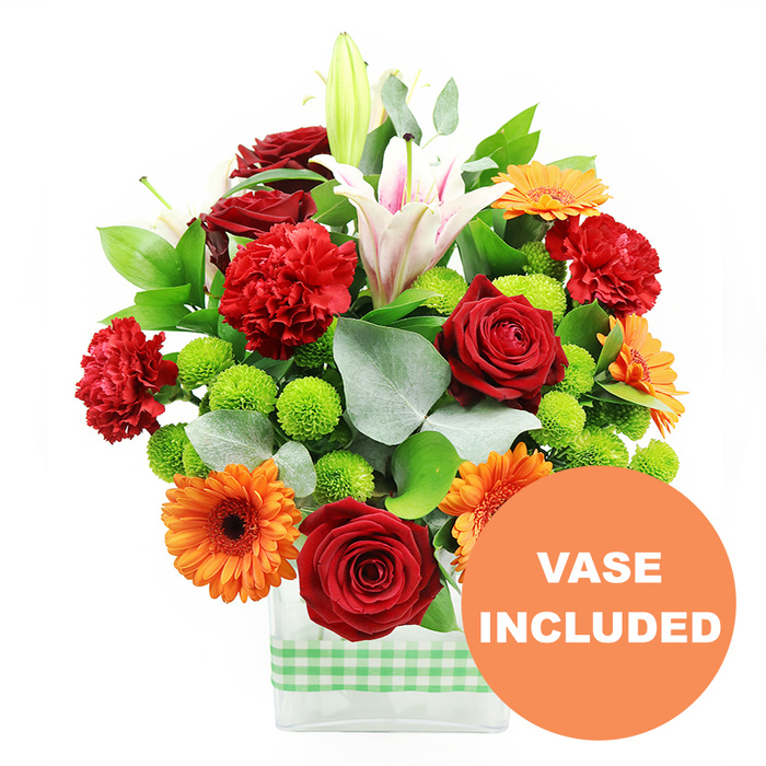This image shows a rich, celebratory bouquet designed with the thoughtful precision of an experienced local florist serving Mill Hill. The arrangement is presented in a modern, rectangular clear glass vase, through which the straight green stems are clearly visible, arranged with care. Around the base of the vase, a light green and white gingham ribbon is tied snugly, giving a soft, rustic accent that feels perfectly at home in the garden-filled streets near Mill Hill Park. Above the vase, the flowers form a lush, rounded mass of colour. Deep red roses, velvety and tightly furled, anchor the composition with a sense of romance and intention. Intermingled with them are ruffled red-orange carnations whose frayed petal edges add texture and movement, making the bouquet appear full and tactile. Bright orange gerbera daisies punctuate the design, their broad, flat petals framing dark central discs, like miniature suns dotted throughout the arrangement. From amidst this warm palette, tall white Oriental lilies emerge, some in full, starry bloom with subtle pink striping along their trumpet-shaped petals, others in smooth green buds that hint at scent yet to be released. Scattered between the larger blooms are clusters of lime-green pompom chrysanthemums, small, rounded, and tightly packed, providing an uplifting, fresh contrast that lightens the overall look. The flowers are supported by glossy, deep green foliage and silver-green eucalyptus leaves, which fan out around the edges, softening the silhouette and adding a cool, calming undertone. Set against a clean white background, the colours appear especially vivid and crisp. In the lower right corner, a circular orange overlay touches the vase, bearing the words VASE INCLUDED in bold white capitals, making clear that this design is delivered as a complete, ready-to-display arrangement, ideal for brightening living rooms, offices, or reception areas across Mill Hill and nearby communities.