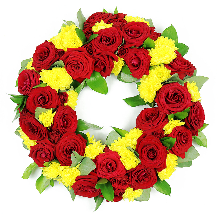 A richly coloured circular wreath is captured here against a plain white background, its design unmistakably intended as a funeral or memorial tribute. The arrangement is dominated by an abundant ring of deep red roses, the blooms pressed closely together so that only glimpses of greenery can be seen between them. Each rose is in fine condition, with gently unfurling petals that range from a classic crimson at the centre to slightly darker, almost burgundy tones near the outer edges, creating visual depth and a sense of luxuriant softness. Interspersed evenly around the ring are clusters of bright yellow chrysanthemums, their many narrow, feathery petals forming round, textured flower heads that bring a vivid, almost sunlit energy to the wreath. These yellow flowers soften the intensity of the red roses and add a sense of warmth and quiet optimism. Supporting the blooms is a base of assorted green foliage, including glossy, medium-sized leaves and lighter, more muted grey-green sprigs that evoke eucalyptus, giving the composition subtle shifts in colour and texture. All elements are arranged with precision to form a smooth, symmetrical circle with an open middle, the classic form symbolising eternal life and enduring remembrance. Bathed in even, bright light, the colours appear pure and saturated, while the textures of petals and leaves are clearly defined. This type of wreath is characteristic of the thoughtful sympathy tributes often ordered in Mill Hill, suitable for dignified services at local churches or crematoria, or for laying at a graveside as a final, eloquent gesture of love and respect.