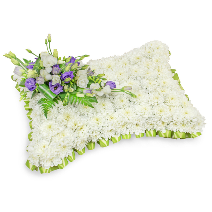 An exquisitely formed memorial pillow, presented against a crisp white studio background, is composed chiefly of hundreds of pristine white chrysanthemums arranged so closely together they read as a single, serene white surface. The small chrysanthemum heads nestle to create a plush, pillow-like silhouette with softly rounded corners and a slightly elevated profile, suggesting comfort and peaceful rest. Around the perimeter a pale lime-green ribbon, pleated into uniform loops, frames the composition with a gentle scallop that lifts the white with a hint of fresh, hopeful colour. Across the upper-left corner an elegant spray of accent florals is placed with considered restraint: velvety white lisianthus are paired with deeper purple lisianthus - some tight as buds, others unfurled - their tones offering a poignant contrast that speaks to respect and remembrance. Interspersed fern fronds and slender green leaves breathe life into the cluster, while a broader dark-green leaf wrap at the stem base secures the grouping with neat, professional finishing. Subtle side lighting emphasizes texture - the chrysanthemum petals' softness, the ribbon's satiny sheen, and the lisianthus' delicate ruffles - evoking quiet moments of reflection you might find during a service near Mill Hill Village or beneath the limes at The Ridgeway. This sympathetic tribute is crafted to convey solace and enduring care, ideal for wakes, funerals, or as a thoughtful graveside offering from neighbours and loved ones in the local area.