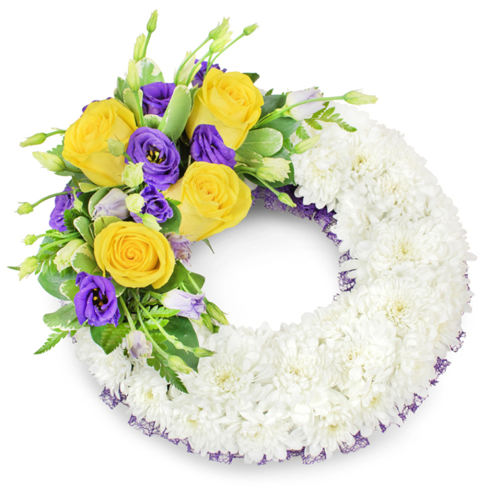 A professionally assembled circular memorial wreath sits composed and serene: the majority of its circumference is densely covered in pristine white spray chrysanthemums, each tiny flower tightly clustered to create a continuous, velvety surface that suggests reverence and unity. From the upper left a concentrated floral accent rises-a measured cluster of bright yellow roses, their layered petals offering warmth and consolation, paired with deep purple lisianthus whose elegant, ruffled blossoms introduce a soft, luxurious contrast. Scattered unopened lisianthus buds in pale lavender-green add depth and a sense of life continuing, while mixed green foliage-shiny oval leaves, delicate fern fronds and lightly variegated stems-frames and supports the colourway. A discreet dark-violet mesh ribbon is visible beneath the white flowers along the inner and outer ring, a refined touch that subtly echoes the purple accents and ties the arrangement together. The wreath's open centre and symmetric form emphasise tradition and the timeless symbolism of the circle. Imagined in soft, neutral studio light, the textures read clearly: the chrysanthemum's cushion-like density, the satin finish of rose petals and the lace-like fronds of greenery. For bereaved families arranging a service in Mill Hill or for deliveries across NW7, this wreath is presented as a thoughtful expression of condolence-simple, composed and crafted with the attention to detail expected from an experienced local florist preparing tributes for funerals or memorial services.