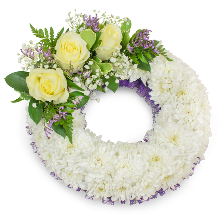 A distinguished round memorial wreath composed primarily of hundreds of small, snow-white chrysanthemums, arranged so densely that the surface reads as a single, plush plane of petals. Each little chrysanthemum head contributes to a tactile, ruffled texture that feels gentle and consoling; the overall tone is pure and reverent. Beneath the white blossoms a refined hint of purple netting or ribbon shows at intervals along the wreath's inner and outer rims, a subtle trimming that adds sophistication and visual depth without intruding on the wreath's serene mood. Anchoring the design in the upper-left sector is a composed cluster of three pale yellow roses-creamy, layered, and fully open-whose soft scent and warm colour offer a note of friendship and solace. These are surrounded by airy clouds of baby's breath (gypsophila) and small sprays of lavender-toned statice, which together create a delicate interplay of textures and a gentle colour counterpoint to the dominant white. Complementary greens-broad, lustrous leaves and feathery fern fronds-provide structure and vitality, framing the flowers and guiding the eye around the circle to the clear, symbolic centre. The wreath sits on a white background under even, natural lighting that produces a soft shadow and highlights the careful craftsmanship. From the perspective of a Mill Hill florist mindful of local memorial rituals near The Ridgeway and Mill Hill Broadway, this wreath is a classic, thoughtful choice that speaks of lasting respect, quiet remembrance, and attention to detail for a funeral or memorial service.