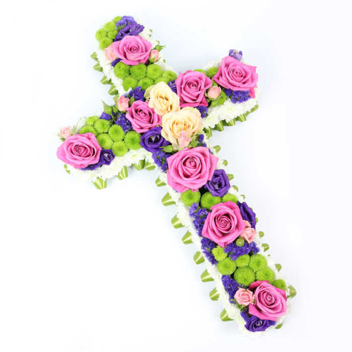 This image shows a beautifully arranged floral cross set against a pure white background, giving the design an almost weightless quality. The cross is slightly tilted, with the upper arm rising towards the left, allowing the viewer to appreciate the fullness and texture of the work. Its outer edge is sharply defined by a fringe of small white spray chrysanthemums, each tiny flower nested closely to the next to form a plush, cushion-like border. Tucked just beneath this layer is a narrow band of soft green ribbon or folded foliage, which runs seamlessly around the entire shape, lending a subtle hint of colour and a finely tailored finish that feels at home in the refined, village-like atmosphere of Mill Hill Broadway. The interior of the cross is densely filled with a carefully balanced mix of blooms. Prominent among them are large roses in vivid fuchsia pink and softer creamy yellow or peach shades, opened fully so that their layered petals form luminous focal points along the arms and stem. Nestled between these roses are clusters of bright lime-green button chrysanthemums, whose small, dome-shaped heads bring lively texture and a contemporary flair. Threaded through the arrangement are smaller flowers in deep purple and violet, probably lisianthus or statice, which provide contrasting shadows and enrich the overall colour story. The craftsmanship suggests patient, attentive hands, placing each stem to ensure both harmony and structure. Illuminated by clear, even light, the cross radiates a blend of solemn reverence and gentle optimism, making it a deeply meaningful sympathy tribute suitable for funerals and memorial services throughout the Mill Hill area.