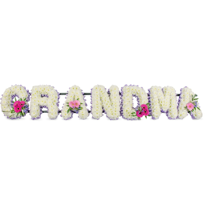 The photograph presents a carefully crafted floral tribute that spells the word GRANDMA in tall, block capitals, each letter fully covered with immaculate white chrysanthemums. The flowers are arranged so closely that their petals overlap in a compact, cushion-like mat, creating the impression of softness and purity. The raised, domed surface of the blooms gives each letter dimension, allowing subtle light and shadow to play across the arrangement. Running neatly around the outer edge of each letter is a fine line of pale lilac ribbon, its satin texture gently reflecting the light. This delicate border traces the shape of the word, adding a calm, refined touch of colour that feels especially appropriate for a grandmother's farewell. Three small posies are placed thoughtfully along the design-on the first G, a central A, and the last A-where clusters of bright pink gerberas and pale pink roses gather with fresh green foliage. These accent sprays provide gentle bursts of colour and organic movement, standing slightly proud from the chrysanthemum base and drawing the eye without disrupting the tribute's serene tone. The arrangement is photographed under soft, even lighting, with a neutral background that keeps attention firmly on the flowers and their textures. It appears as if it has just been finished by a skilled florist in Mill Hill, perhaps in a studio not far from Mill Hill Park, and made ready for delivery to a local funeral service or chapel of rest. The overall feeling is one of quiet affection and gratitude, a floral statement that plainly acknowledges the word GRANDMA while also carrying generations of family stories, Sunday lunches, and gentle encouragement within its petals.