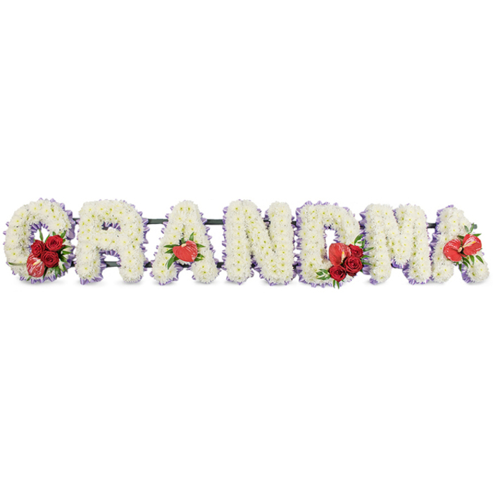 The photograph depicts a loving funeral tribute in the form of floral lettering spelling out GRANDMA, thoughtfully arranged as if to be placed in front of a coffin or near a memorial table in Mill Hill. Each letter is sculpted from dense clusters of white cushion chrysanthemums, whose uniform, rounded blooms create a velvety, almost quilted effect across the surface. There is a subtle halo of pale lavender or light purple ribbon or backing edging every letter, just visible beneath the petals, lending a soft, pastel outline that helps the word stand crisply against the plain white background. Into this serene backdrop of white and lilac, the florist has woven intense red highlights that symbolise affection and remembrance. The opening G features a cluster of deep red roses nestled into its curve, paired with a distinctive red-and-white variegated calla lily, its graceful arc and delicately patterned petals providing a striking contrast. The first A is adorned with a single heart-shaped red anthurium blossom at its base, its smooth, waxy surface and prominent spadix adding texture and a contemporary touch. At the centre of the tribute, the M is enriched with another grouping of red roses and a matching red anthurium, creating a natural focal point; the last A echoes this with a final red anthurium bloom, offering balance to the composition. The letters are quietly supported by a dark green framework, almost hidden from view, ensuring the tribute can be displayed securely at a Mill Hill funeral home, church, or cemetery. The design reflects skilled craftsmanship and heartfelt intention, providing a graceful way for a Mill Hill family to honour their grandmother's life, blending tranquillity and warmth in every petal.