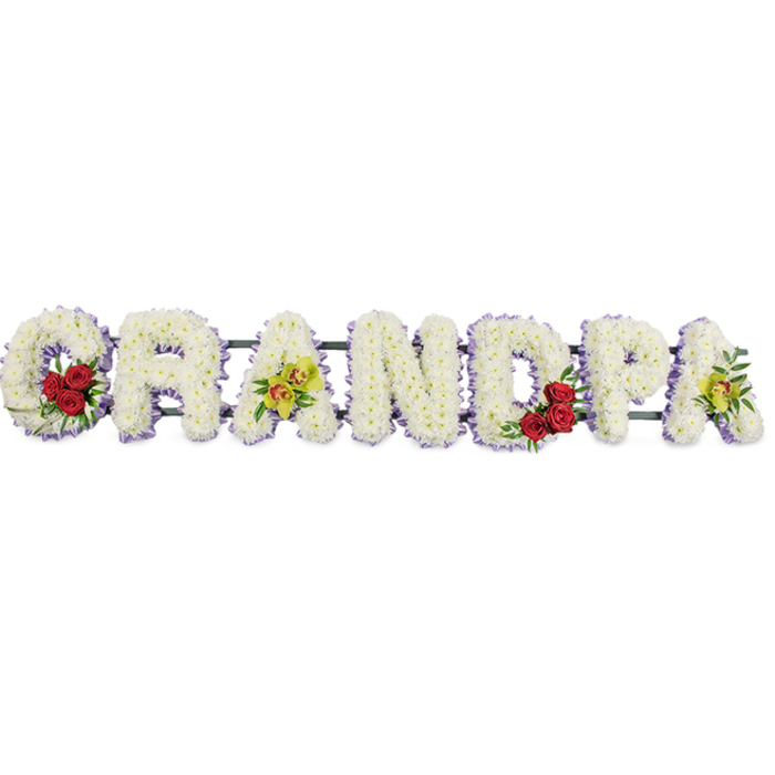 A beautifully composed sympathy tribute stretches across the frame, spelling out GRANDPA in bold, block capital letters, each one standing out crisply against a plain white background. The florist has meticulously packed the letters with pristine white chrysanthemums, arranging the small, rounded flowers so closely that they form a thick, textured blanket of petals. This dense covering gives the impression of softness and quiet comfort, as though the flowers were gently supporting the memory of a much-loved grandparent. Tracing the outer edge of every letter is a fine line of lavender-purple ribbon, which softly defines the shapes and adds a hint of colour reminiscent of evening light over the rooftops of Mill Hill. Carefully chosen accent blooms introduce warmth and individuality. In the sweeping curve of the first G, three to four deep red roses gather in a rich cluster, surrounded by fresh green foliage that sets off their velvety petals. The third letter, A, is decorated with refined yellow-green orchid blossoms, some touched with a delicate reddish hue, lending the design a quietly luxurious air. The curved D echoes the earlier G with another cluster of red roses, while the final A repeats the orchid spray, visually tying the tribute together from one end to the other. Behind the letters, slim horizontal bars are just visible, discreetly holding the structure steady so it can be displayed at a funeral in Mill Hill, perhaps in a local church off Daws Lane or near the open spaces of Arrandene. Crafted by Flowers Mill Hill with sensitivity and skill, this arrangement blends serene white, romantic red, and graceful orchid tones to express love, gratitude, and peaceful farewell for a grandfather.