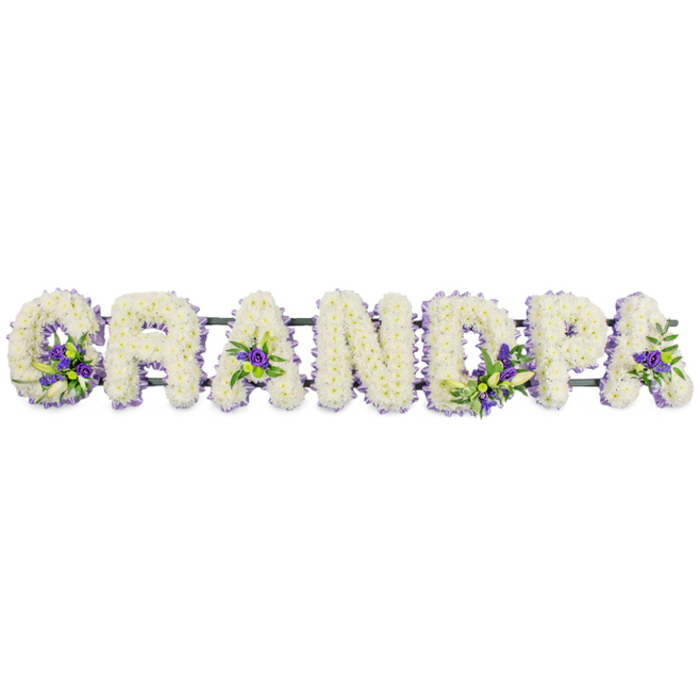 The scene captures a personalised funeral flower arrangement spelling GRANDPA in large, evenly spaced capital letters, displayed horizontally as you might see in front of a coffin or on a stand at a service in Mill Hill. Each letter is densely upholstered with small, pristine white chrysanthemums or carnations, creating a thick, plush layer of petals that looks almost like soft fabric from a distance. The blossoms are chosen for their longevity and neat, rounded form, giving the entire tribute a polished, composed appearance. The pure white colour radiates a sense of innocence, reverence, and quiet respect, perfectly suited to a final farewell. To soften the expanse of white and introduce gentle depth, the florist has added carefully arranged clusters of purple and lavender blooms, possibly lisianthus or small chrysanthemums, interlaced with sprigs of fresh green foliage. These accents are placed with deliberation: a prominent grouping at the base of the G, a tucked-in spray within the triangle of the first A, a touch of colour in the upright of the D, more blossoms along the P, and a final accent on the last A. These spots of purple draw a delicate visual path across the word and echo traditional mourning colours. Beneath the flowers, a dark green structural bar holds the letters in place, mostly concealed by the lush covering of petals but vital for maintaining their alignment during transport and display. Set against a clean, neutral background, the tribute feels serene and contemplative, reminiscent of quiet corners in local Mill Hill churches or the peaceful surroundings near Mill Hill Park. It serves as a meaningful, customised expression of love and remembrance for a grandfather, transforming grief into a beautiful, dignified floral statement.