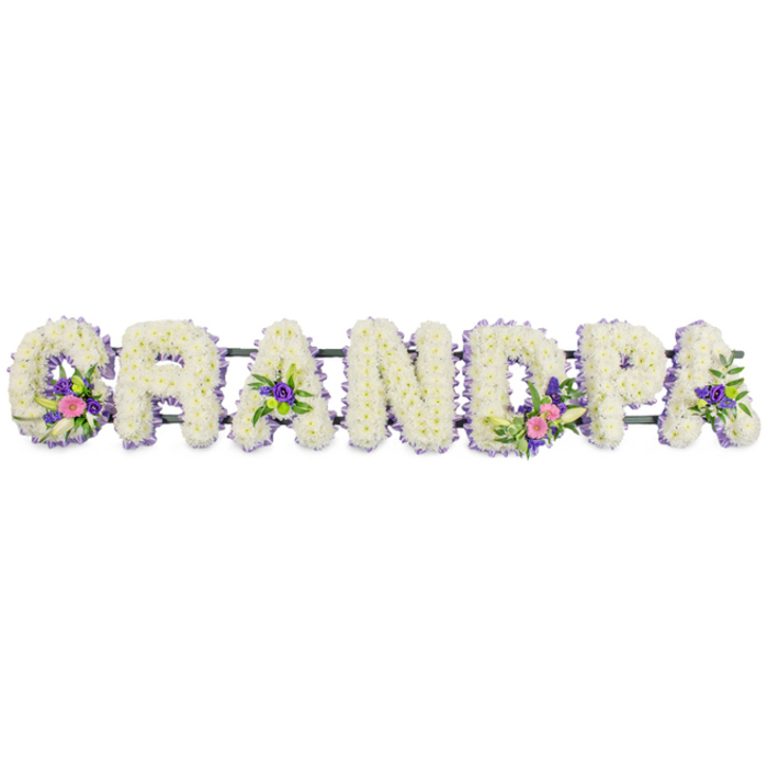 This photograph shows an elegant, personalised funeral flower arrangement spelling out GRANDPA in large, capital letters, crafted with the kind of precision and care you might expect from Flowers Mill Hill for a service at a local church or crematorium. The seven letters are aligned in a neat horizontal row, each one completely covered with small, pristine white chrysanthemums or similar multi-petaled flowers, layered densely to create a rich, cushion-like texture that feels soft and consoling. Around the perimeter of every letter runs a distinct but understated border of tiny lilac and pale purple blooms, probably statice or heather, forming a fine line of colour that enhances the legibility of the word while maintaining a gentle, respectful palette. Within selected letters, accent sprays bring movement and subtle colour contrast: the G carries a cluster of pink and fuchsia flowers, along with an elegant, star-shaped white lily-like bloom and fresh green foliage that arches lightly over the white base. The first and final A include smaller groupings of deep purple florets and glossy leaves, while the D features a more pronounced cluster echoing the G with vivid pinks, white petals, and vibrant greenery; hints of bright green button chrysanthemums peek through, adding another layer of texture and freshness. All the letters are fixed to a slender, dark green metal frame running discreetly behind them, ensuring the tribute can be displayed securely and visibly. Photographed against an immaculate white background with soft, even lighting, the arrangement exudes calm and dignity. Each carefully placed stem seems to speak of fond memories, gentle humour, and the steady presence of a much-loved grandad from the Mill Hill community.