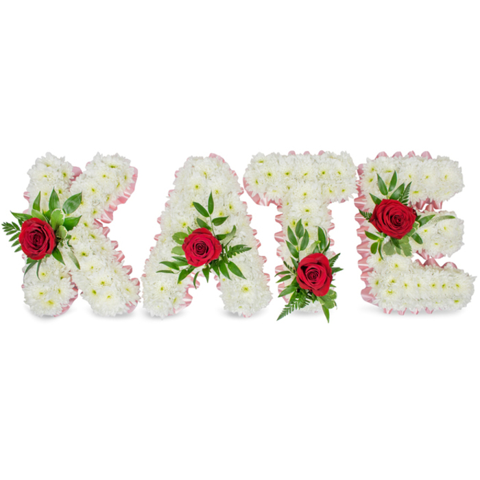A beautifully composed floral name tribute reads KATE in large, sculpted letters formed entirely from densely packed white chrysanthemums. The small, pristine chrysanthemum heads create a rounded, pillowy surface across each letter, producing a serene, cloud-like silhouette that denotes purity and gentle reverence. Each letter's outer contour is trimmed in a light pink satin ribbon, the pleated ribbon edging introducing a soft, warm outline that lifts the composition from the white background. On the face of every letter a solitary red rose provides a vivid point of attention - their deep crimson petals stand out against the immaculate white, while their velvety texture contrasts with the matte chrysanthemums. The roses are embraced by a generous spray of foliage: glossy broad leaves and fine fern-like fronds combine to add depth, natural rhythm and a sense of quiet movement. Attention to placement is clear and considered: the lower portion of the left vertical stroke of the K holds its rose and greenery, the A's rose crowns the apex of its triangular form, the T is balanced by a rose at the base of the left side of its crossbar, and the E is decorated with a rose on its central horizontal bar. The arrangement sits on a clean white backdrop with soft, even light that highlights form and shadow without distraction. Suitable for funeral services, memorial displays or a commemorative gathering in Mill Hill Park or nearby chapels, this personalised floral lettering expresses respect, love and lasting memory with classic colour harmony.