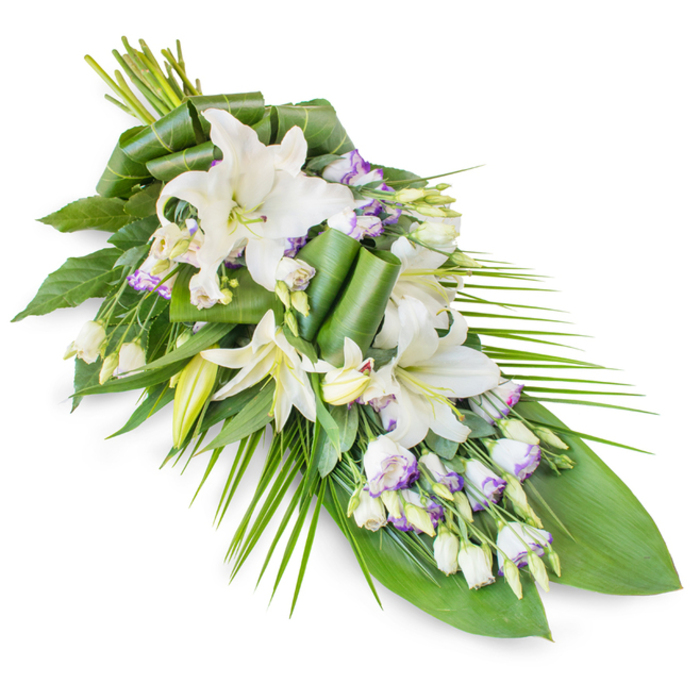 A graceful memorial flower spray is displayed against a clean white backdrop, its design flowing diagonally like a quiet stream of petals and leaves. The arrangement begins with a dense, rounded gathering of stems and blooms at the upper left and gently tapers to a fine, pointed tip towards the lower right, reminiscent of a traditional teardrop or casket spray. At its core are white oriental lilies, which command attention with their large, open blooms and satin-like petals. Some lilies are fully unfurled, their pale yellow-green stamens and gently curved petals clearly visible, while others remain in elongated buds, capturing a sense of pause and reflection. Threaded throughout the lilies are clusters of lisianthus, predominantly white blossoms traced with delicate lavender edges and fine purple veining that echo the soft twilight tones over Mill Hill's green spaces and quiet avenues. Numerous tight lisianthus buds contribute height, texture, and a sense of continuity, as though the tribute is still gently coming into being. The surrounding foliage is rich and varied: broad, dark green leaves provide a strong, grounding base, while slimmer, palm-like fronds fan outward, especially on the right side, lending movement and elegance. Some leaves are precisely folded, looped, and rolled, forming modern, architectural shapes that reveal the hand of an experienced florist used to preparing tributes for Mill Hill churches, crematoriums, and family gatherings. The overall effect is serene and dignified, with the interplay of white blooms and deep green foliage suggesting purity, peace, and the enduring love that holds a family together. This spray would sit beautifully atop a casket or as a central display at a memorial service, offering quiet comfort and respect.